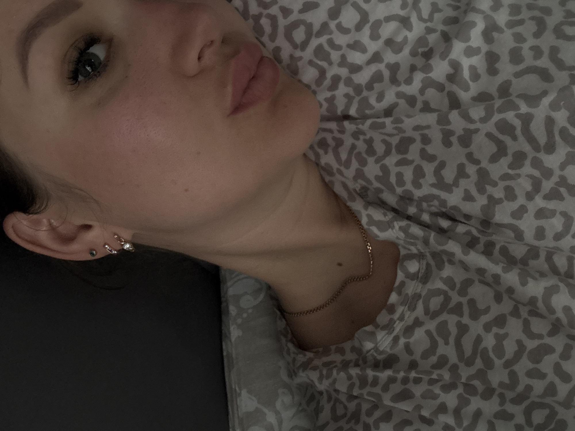 A close-up, slightly rotated, indoor photograph captures a person from the chest up. The person has fair skin, light brown hair, and notable features include full lips, defined eyebrows, and eyelashes enhanced with mascara. Two earrings are visible in the left ear, and a delicate gold chain necklace rests on the neck. The person is lying on a surface covered with a gray and white animal print fabric, likely a bed or a couch. The background is a dark, undefined surface, possibly a wall or furniture. There is no visible information indicating the city of Gelendzhik, Russia.