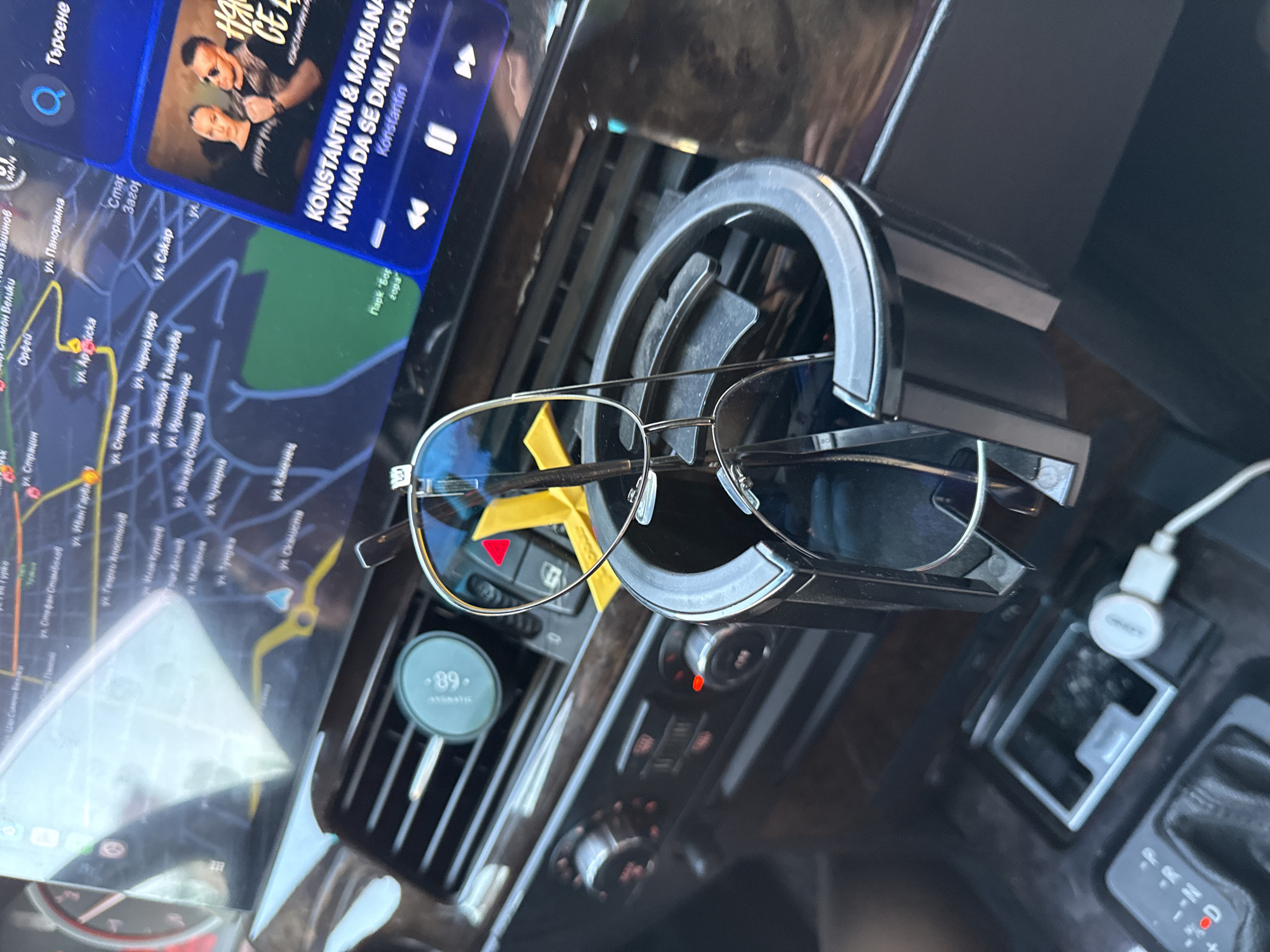 An elevated-angle, interior shot depicts the dashboard and center console of a vehicle, likely a BMW due to visible design cues. A large display screen dominates the upper portion, bifurcated into a navigation map on the left and a music player interface on the right. The map, rendered in Cyrillic, displays street names such as "Ул. Стефан Стамболов," "Ул. Цар Симеон Велики," and "Ул. Иван Гарба," with a prominent label identifying "Стaра Загора," locating the scene in Stara Zagora, Bulgaria. A green park area, labeled "Парк "Бел еopa"," is also visible. The music player displays album art featuring a man and a woman, with the text "КОНСТАНТИН & MARIANA" and the song title "НЯМА ДА СЕ ДАМ / КОН..." (NYAMA DA SE DAM), attributed to "Konstantin." A search icon and the word "Търсене" (Search) are visible in the upper right.
Below the screen, a dark wood-grain or similar panel runs across the dashboard. Air conditioning vents are visible, with a circular dark green device clipped to one, displaying "89" and "aromatica." To the right, a black plastic cup holder insert contains two pairs of aviator-style sunglasses: one with blue-tinted lenses and another with darker lenses, both suspended or resting within the holder. A red triangular warning light is partially visible behind the sunglasses. Below the vents, three rotary climate control knobs are present, two of which have illuminated red indicators. The automatic gear shifter, displaying "P," "R," "N," and "D," has the "D" (Drive) indicator illuminated in red. A white USB charging cable is plugged into a port positioned in a small storage compartment adjacent to the gear shifter. No individuals are depicted in the image.