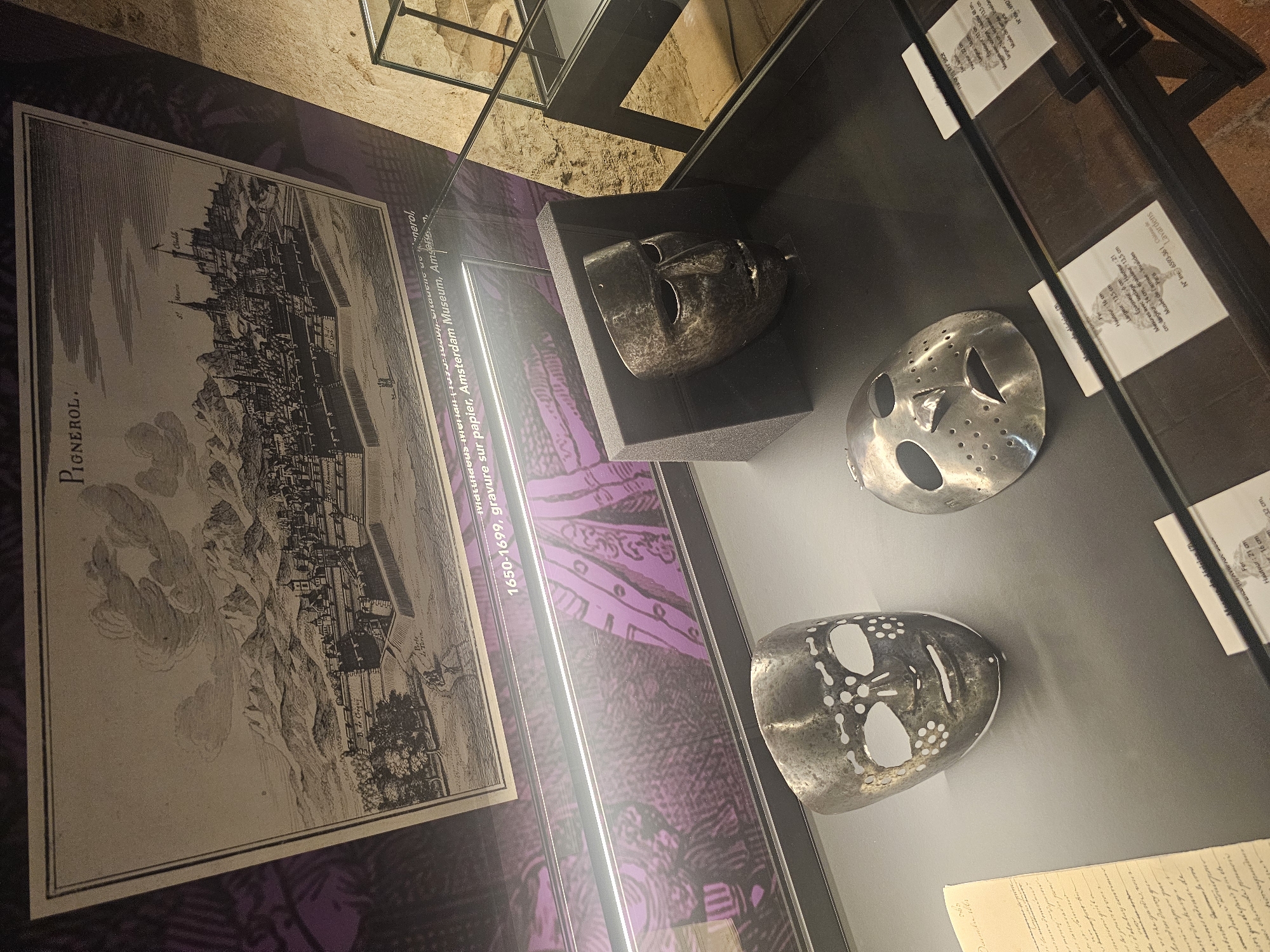 This image shows a museum display case containing three metal masks and a historical engraving. The engraving, titled "PIGNEROL," depicts a walled city with buildings and mountains in the background. Text below the engraving reads "1650-1699, gravure sur papier, Amsterdam Museum, Amsterdam." Inside the display case, two of the masks are positioned on the left, with one slightly in front of the other. The mask on the left has numerous small circular perforations across its surface, and large, roughly oval eye openings. The mask in the middle is less perforated, with fewer holes around the mouth and nose area, and is positioned closer to the viewer. A third mask, darker and more primitive in appearance, sits on a black stand on the right side of the display. A handwritten document is visible to the left of the masks. The background of the display features a dark, patterned fabric and a stone wall.