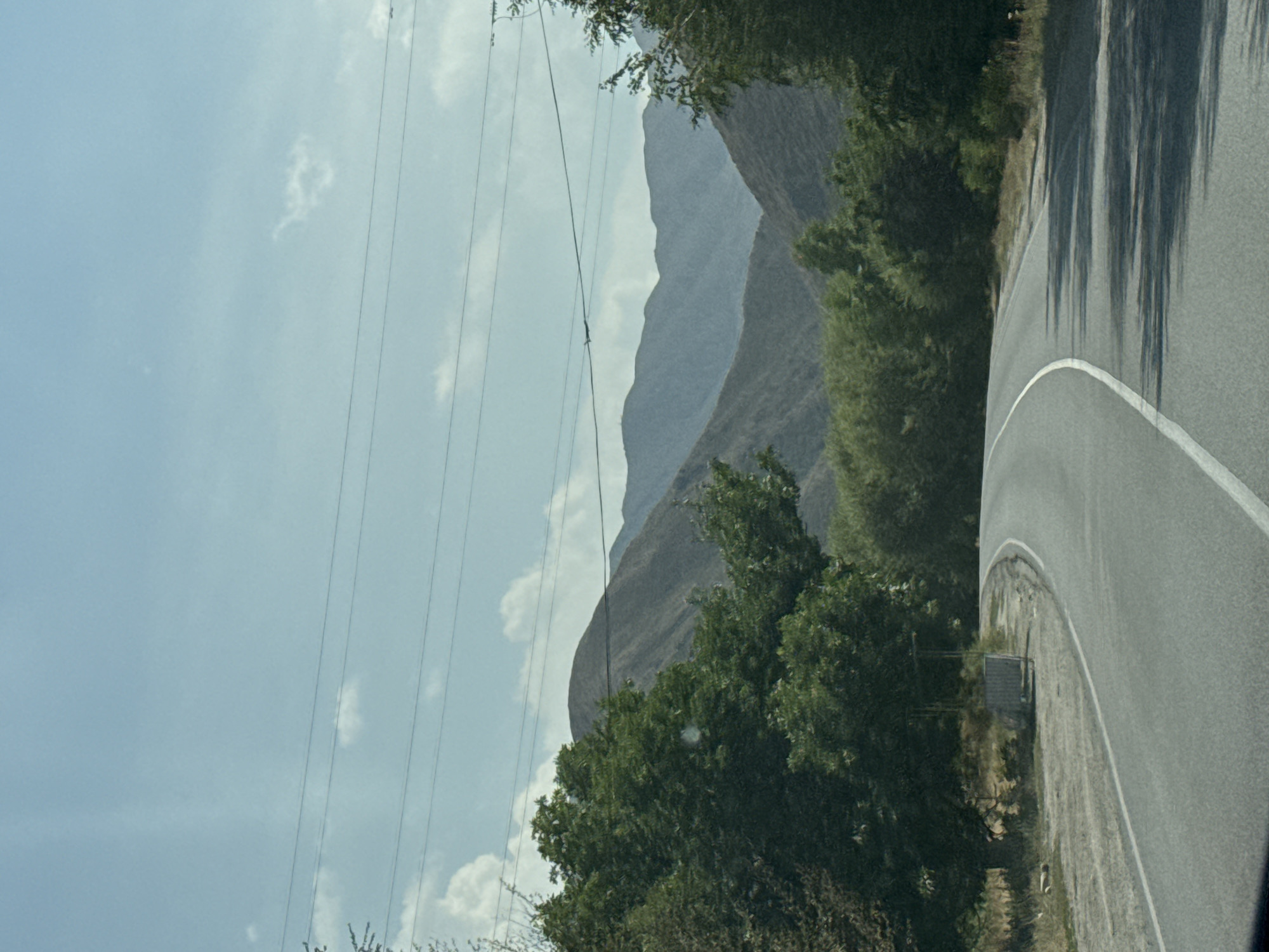 The image shows a paved road curving to the right, leading through a landscape with trees and distant mountains. The sky is clear with some scattered clouds. Power lines are visible stretching across the sky. In the foreground, the road has a white line marking its edge. To the left of the road, there are some low bushes and a small structure that appears to be a bus stop shelter. The mountains in the background are layered and appear arid. The overall scene suggests a rural or semi-rural setting. The image was taken in the vicinity of Koi-Tash, Kyrgyzstan.