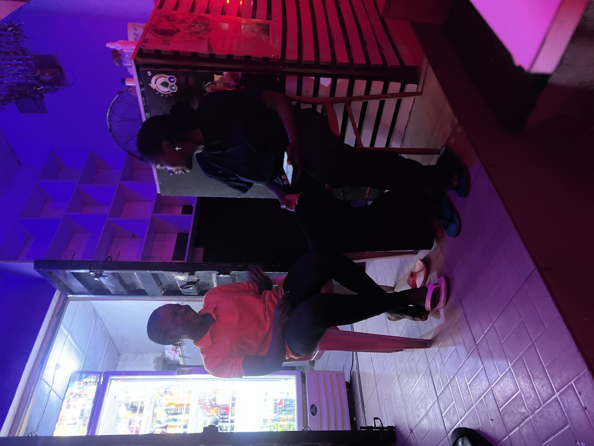 This image captures two individuals in a casually lit indoor setting in Howayi, Nigeria.
On the left, a person with short dark hair, wearing an orange polo shirt and dark pants, is seated on a light pink plastic chair. This individual is smiling broadly and looking towards the right, with hands slightly gestured as if mid-conversation. Bare feet are visible, with one foot partially in a pink flip-flop. Behind this person, a bright white-lit area reveals a tall, glass-front commercial beverage refrigerator filled with various bottled and canned drinks. A white wall and a ceiling light are also visible in this area.
On the right, a second person with dark hair, wearing a dark (possibly black) t-shirt and dark pants, is seated on a similar chair, partially obscured by a foreground element. This individual holds a smartphone with a glowing screen in both hands and is looking towards the left, engaging with the first person. A small silver cross earring is visible in their left ear. Blue flip-flops are on their feet. To the right of this person is a structure with horizontal dark and light stripes, illuminated by red strip lighting. Above this structure, a black fan and some decorative tinsel-like elements are visible on the ceiling. A piece of dark equipment with a white skull-like logo is present on a surface behind the second person.
The overall environment is characterized by distinct lighting zones: a dominant purple-blue hue illuminates most of the space, contrasting with the bright white light from the refrigerator area and the red light emanating from the right-side structure. The floor consists of light-colored square tiles, reflecting the varied lighting. Both individuals appear relaxed and engaged in a friendly interaction.