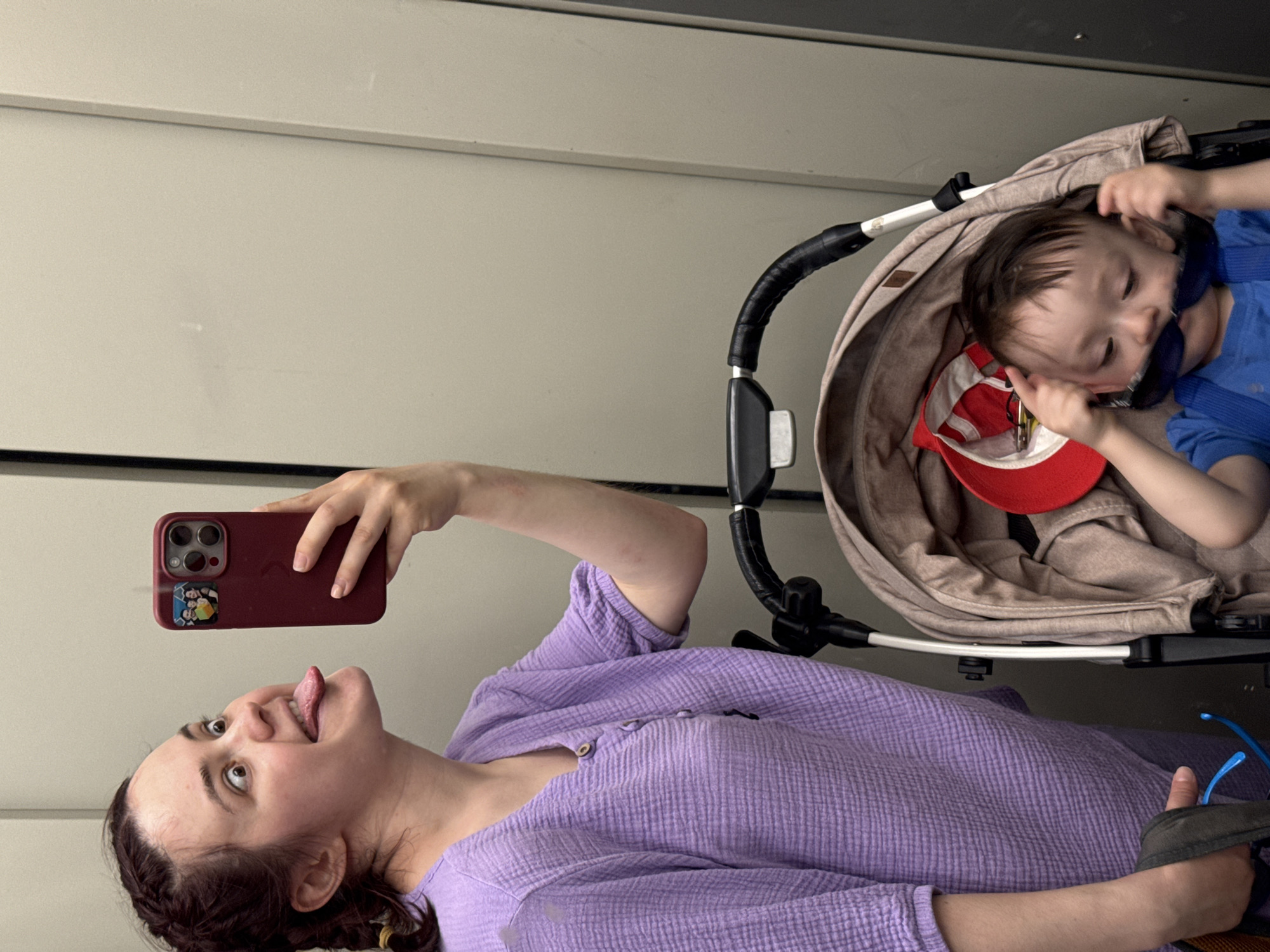 A woman with braided hair and a purple shirt is taking a selfie with her phone, sticking her tongue out. A child is in a stroller beside her, wearing sunglasses and a blue shirt. A red baseball cap is inside the stroller next to the child. The location is indeterminate, and there is no evidence to suggest the scene is in Izhevsk, Russia.