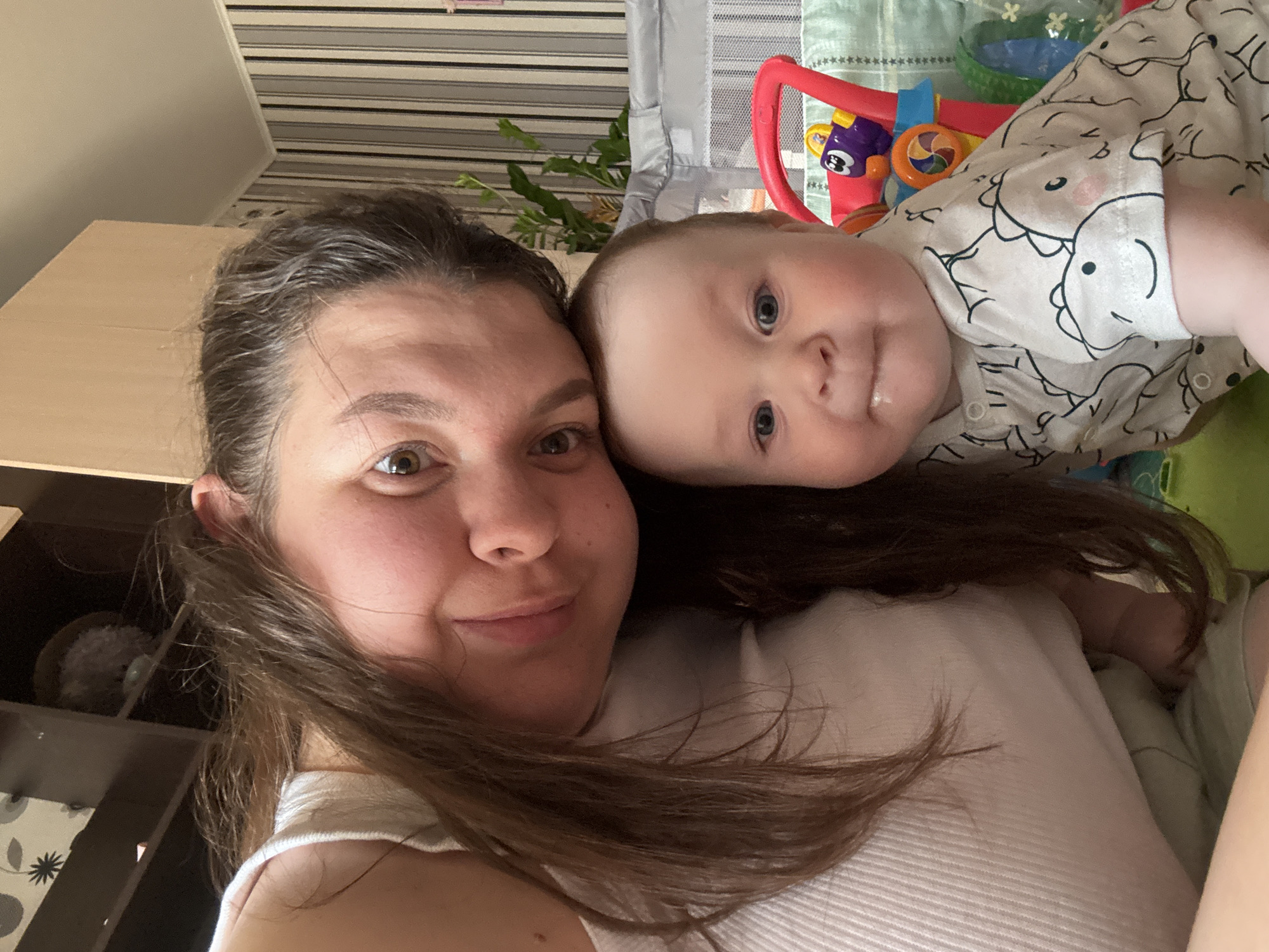 A selfie photograph shows a woman and a baby lying on a light-colored, horizontally striped surface, likely a bed. The woman has brown hair and is wearing a white top. The baby, wearing a white onesie with a dinosaur print, is lying close to the woman's head. In the background are stripes and a green patterned blanket, a red toy, and a green basket. A piece of furniture is partially visible in the background, suggesting the setting is indoors. The image was likely taken in the city of Марковский, Russia, given the request.