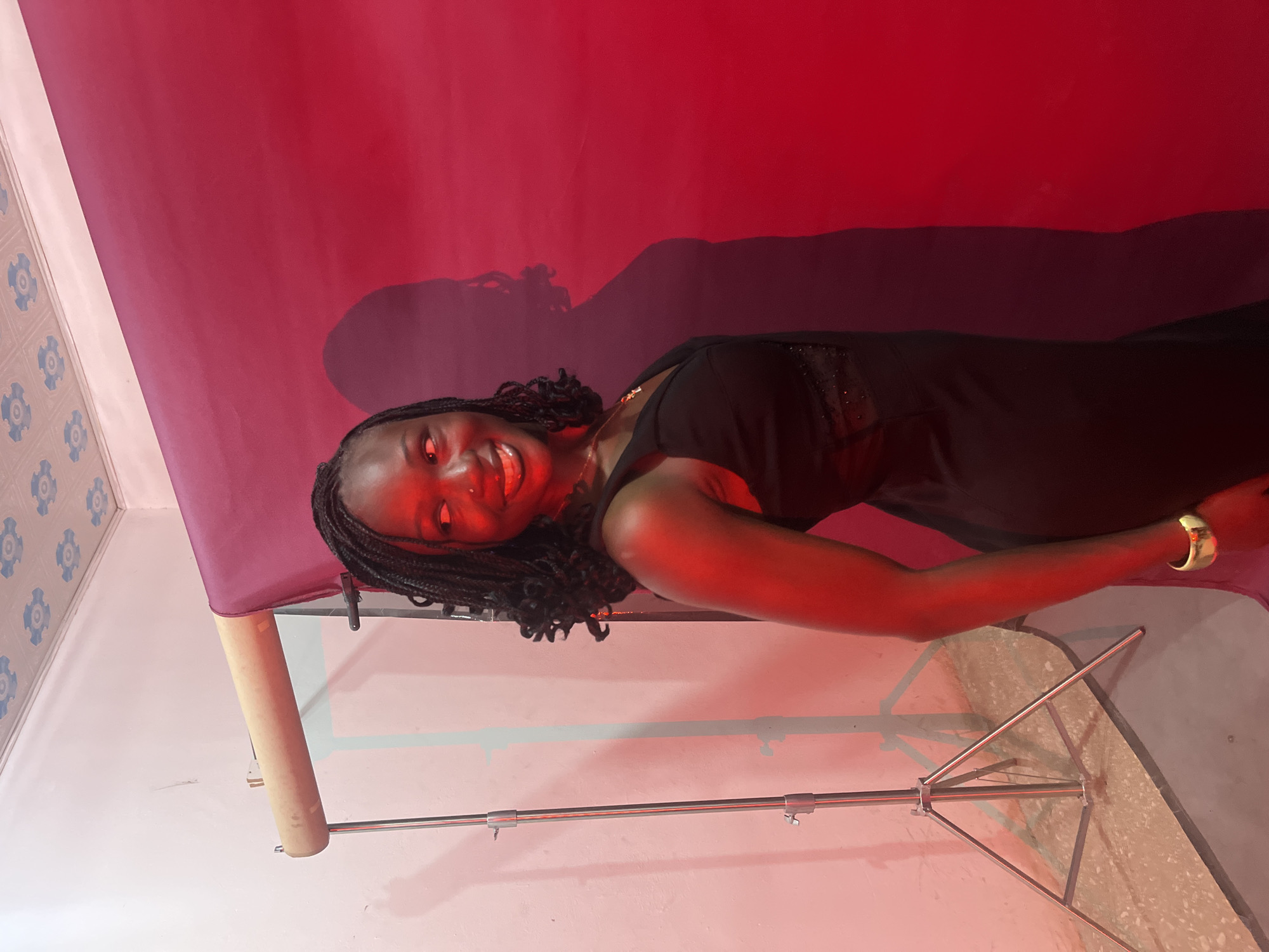 A young Black woman with braided hair is posing for a photograph against a red backdrop in a studio setting. She is wearing a black sleeveless dress with a deep V-neckline and decorative detailing on the bodice. A golden bracelet adorns her left wrist. The lighting is predominantly red, casting a strong glow on her skin and creating a prominent shadow of her silhouette on the backdrop. To her left, a photography lighting stand is visible, along with a pale pink wall and a section of the studio ceiling with a floral pattern. The floor appears to be a textured concrete or tiled surface. The scene is likely within a photography studio in Gura Top, Nigeria, as indicated by the provided context, though this specific location cannot be definitively confirmed from the visual information alone.