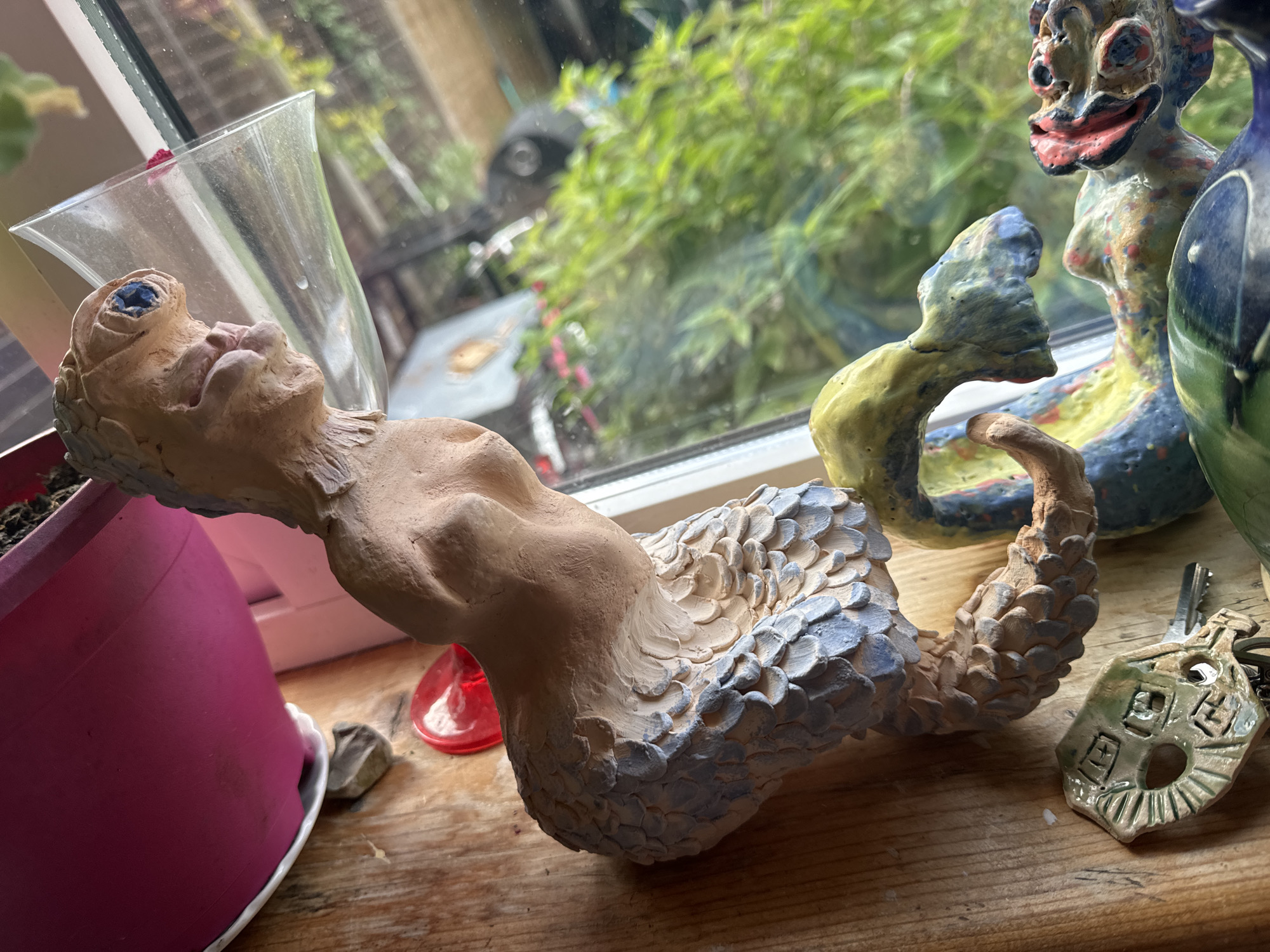 A close-up, eye-level shot captures a detailed view of several whimsical ceramic figurines and a pink planter on a wooden surface, illuminated by natural light filtering through a window. The figurines are rendered in a folk art style, with uneven textures and vibrant, varied color palettes.
The most prominent figurine, positioned in the foreground, depicts a creature with a humanoid torso and a tail resembling fish scales. The scales are textured and painted with shades of light blue and beige. The creature's head is tilted back, with large, bulbous eyes, and its mouth is slightly open, revealing pink inner lips. It rests near a clear, trumpet-shaped glass vase and a bright red, heart-shaped object.
Behind this figure, another ceramic figurine stands tall, with a more grotesque yet charming expression. This figure is painted with a mosaic of colors, including blues, yellows, reds, and greens. Its body is elongated and appears to be in a dance-like pose. Partially visible to its right is the rim of a green and blue glazed vase.
In the lower left of the frame, a bright pink planter holds what appears to be a plant, though only the edge of a green leaf is visible. To the right of the main figurine, a keychain with a ceramic charm shaped like a house, featuring windows and a door, rests on the wooden surface. A metal key is attached to the charm.
The background is dominated by a softly blurred view through a window, revealing lush green foliage and hints of what might be outdoor furniture or structures. The overall impression is one of a vibrant, artistic display set on a rustic wooden surface, likely a windowsill or shelf. The scene is not identifiable as being located in Waterford, Ireland.