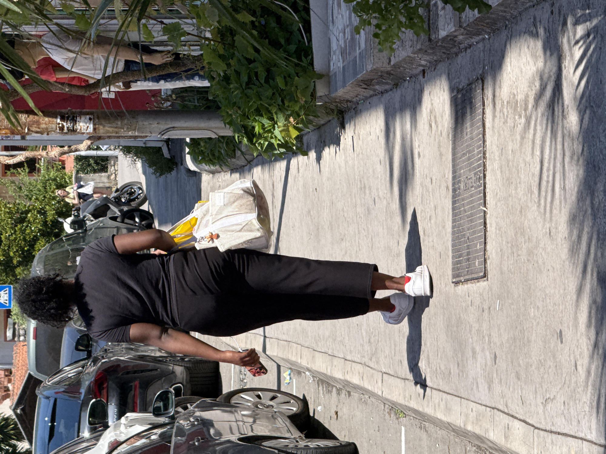 In Budva, Montenegro, a Black woman walks away from the camera on a sunlit sidewalk. She wears a black t-shirt and black pants, and white sneakers with red accents. She carries a large, light-colored tote bag in her right hand and a cell phone in her left. Parked cars line the street to her left, and a building with a red awning and greenery is visible on her right. The scene appears to be a commercial or residential street.