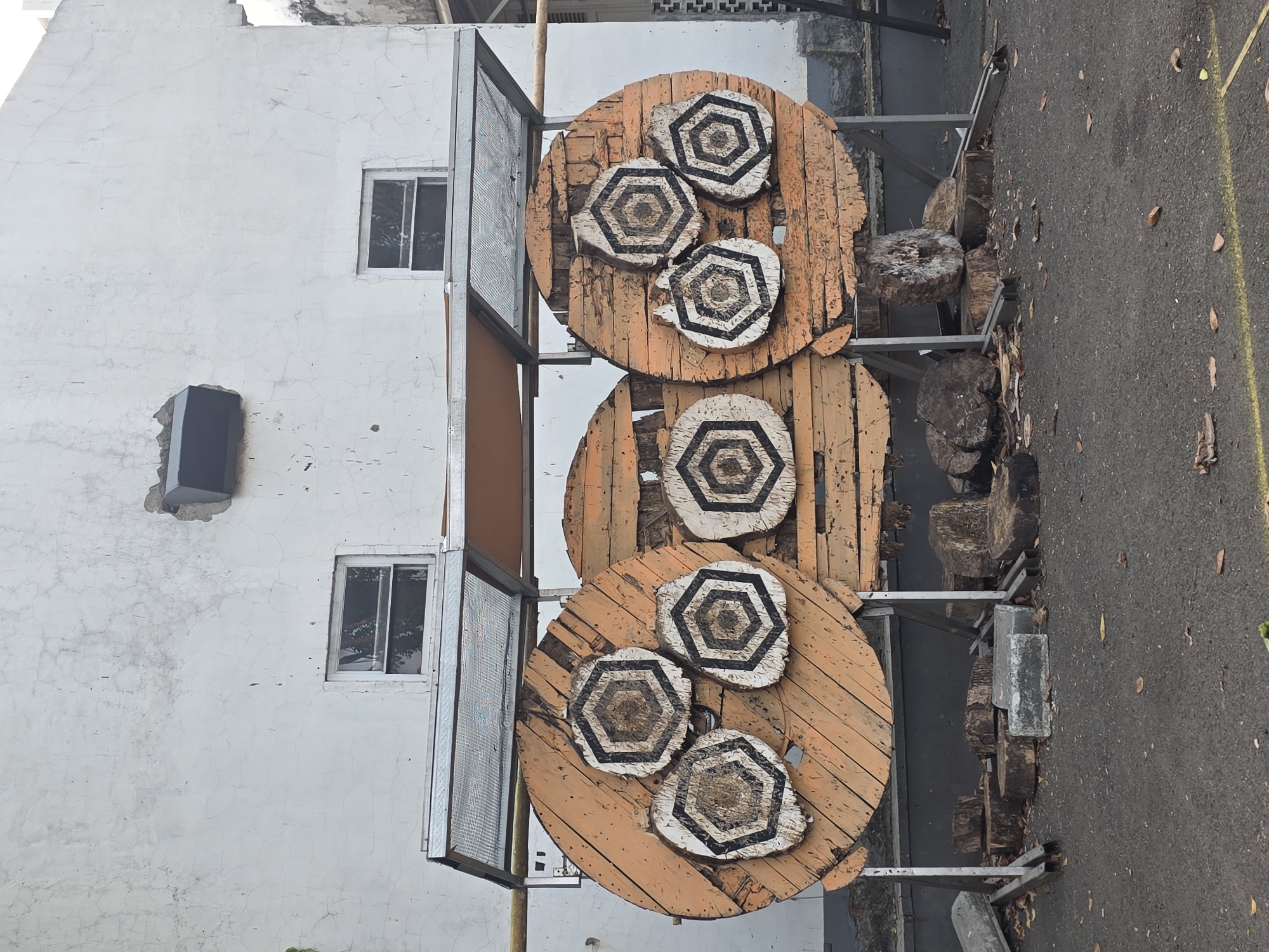 The image shows an outdoor axe-throwing range in Tambun, Indonesia. Three large, circular wooden targets are set up in front of a white, weathered building with two small windows. Each target is made of several planks of wood and has smaller hexagonal targets attached to its surface. These smaller targets feature a geometric pattern of black and white rings. The targets are supported by metal stands. Scattered around the base of the targets are several large wooden stumps, some of which are weathered and split. The ground is paved with asphalt, and fallen leaves are visible on the ground. A metal roof structure extends over the targets.