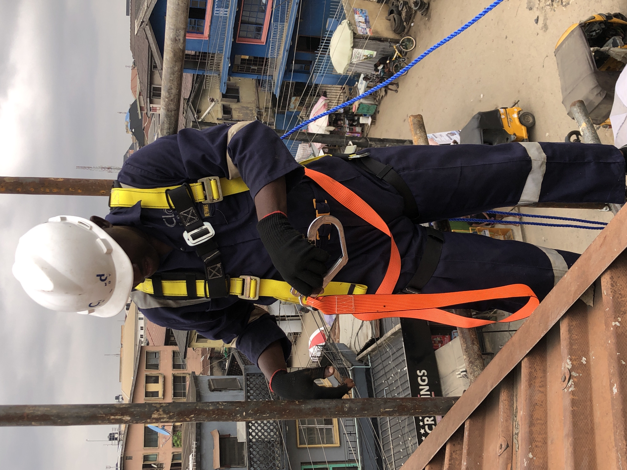 The image captures a construction worker, wearing a white hard hat, dark blue overalls, and a bright yellow safety harness, on a rooftop in Ikeja, Nigeria. The worker is holding an orange safety lanyard with a metal carabiner. The background reveals a dense urban landscape with buildings of various colors and heights, characteristic of Ikeja. Power lines crisscross the scene, and several yellow auto-rickshaws, known as "Keke NAPEPs," are visible on the street below. The sky is overcast, suggesting a cloudy day. The overall impression is one of a typical working day in a bustling Nigerian city, with a focus on the safety precautions being taken by the worker.