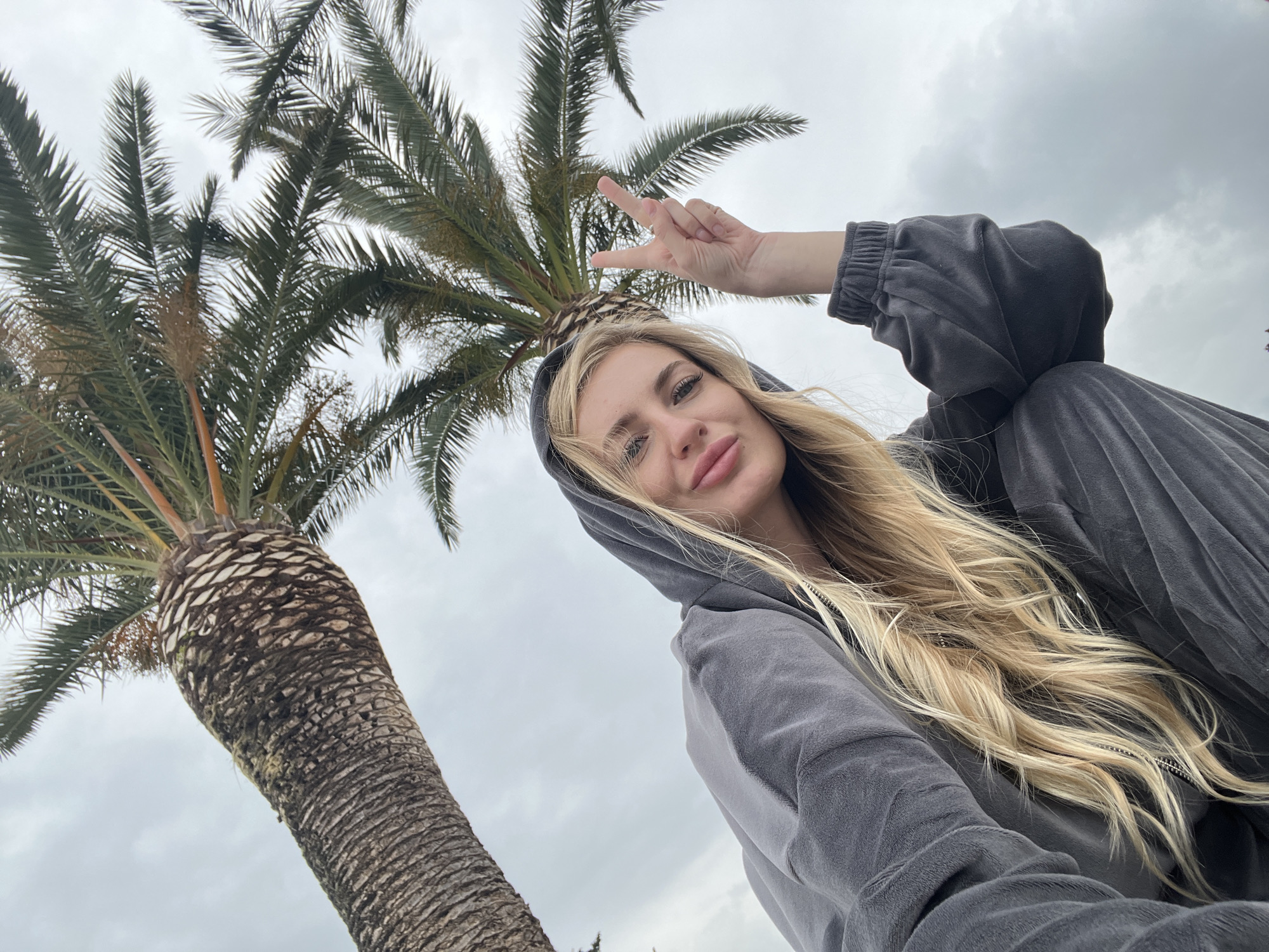 A low-angle, medium shot depicts a young woman in a gray hooded sweatshirt, making a peace sign with her right hand. Her blonde hair is long and flows down past her shoulder. Behind her is a palm tree with a thick trunk and full canopy. The background is an overcast sky. The location is most likely Budva, Montenegro, considering the palm tree and the individual's likely geographic location based on the provided information.
