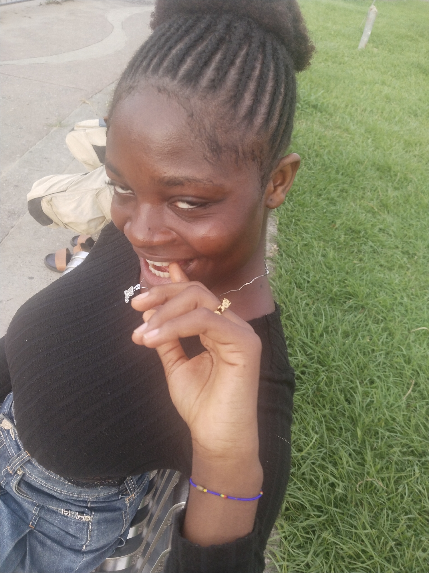 A medium, close-up shot captures a young woman of African descent outdoors, likely in Uyo, Nigeria, judging by the subject's appearance. She has dark brown skin, braided hair styled up into a bun, and is wearing a black, long-sleeved shirt and denim jeans. The woman is smiling slyly and has her index finger partially in her mouth. She is also wearing a silver necklace with a charm and a beaded bracelet on her left wrist.
Behind the woman is a backdrop of green grass and a concrete area, potentially a sidewalk or curb. A bag and footwear are also visible in the background. The scene is well-lit, suggesting daytime.