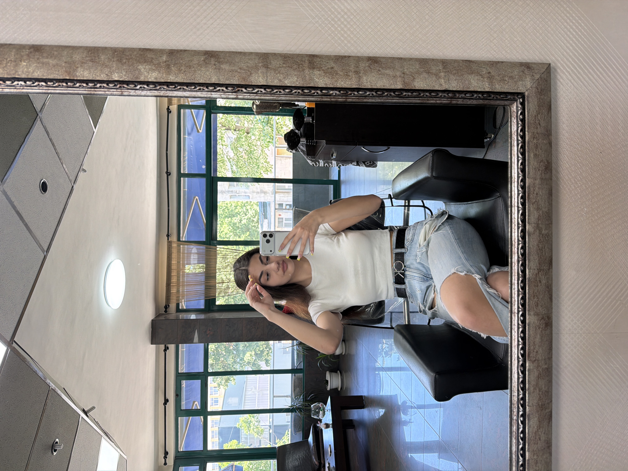A mirror reflects a young woman taking a selfie in what appears to be a hair salon or beauty parlor. She is seated in a black styling chair, wearing a white shirt and ripped blue jeans, with a black belt. The reflection shows a room with large windows overlooking a city scene with buildings and trees, possibly in Gabrovo, Bulgaria. Other furniture and salon equipment are visible in the reflection.