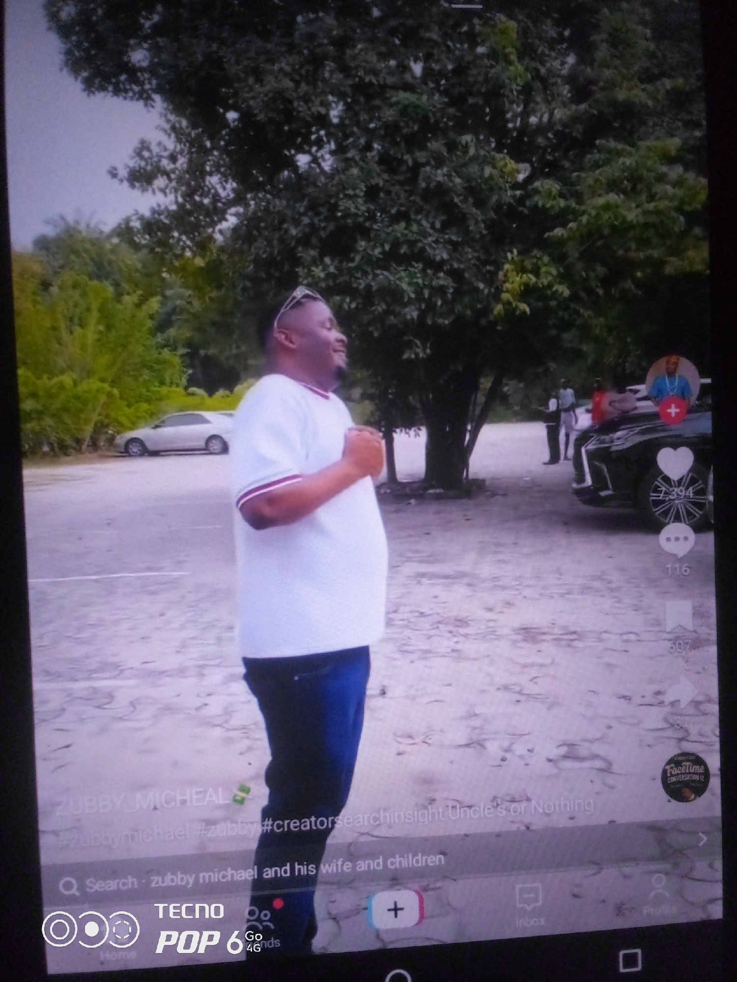 In the city of Howayi, Nigeria, a man in a white t-shirt and blue jeans stands facing right, with his hands clasped in front of him. He is positioned on a sandy, outdoor surface, with trees and foliage forming a natural backdrop. In the background, a silver sedan is parked on the left, and a black Lexus SUV is visible on the right, with a few people standing near it. The scene appears to be a casual outdoor gathering or event. The image is a screenshot from a mobile device, indicated by the on-screen interface elements such as hashtags and icons for search, likes, and comments.