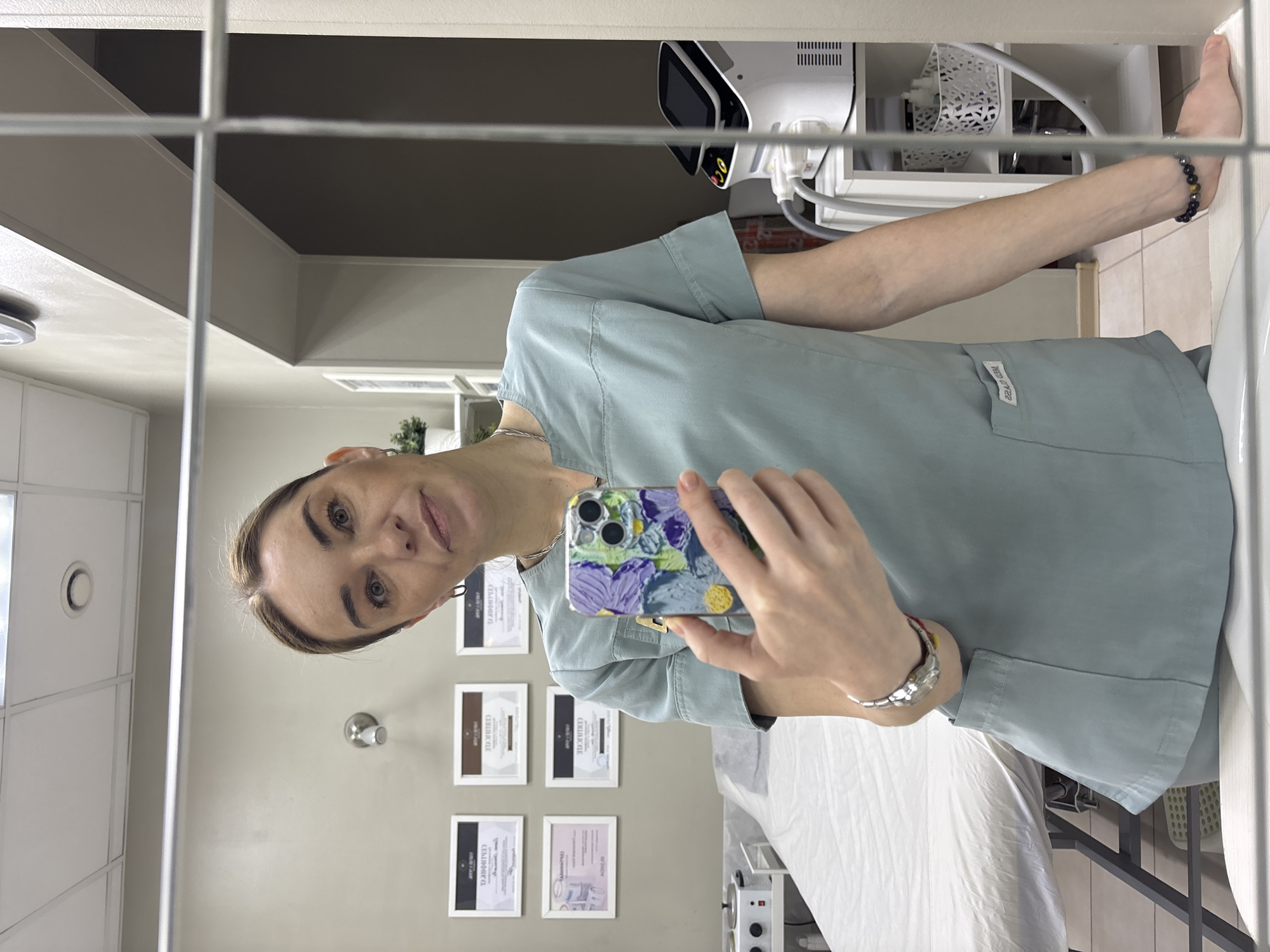 A woman in a light blue scrub top is taking a selfie in a mirror. She is holding a cell phone with a floral-patterned case. Her left arm is extended and wears a bracelet. The background includes a treatment bed with a white sheet, framed certificates on the wall, and a medical device with a display screen on a shelf.