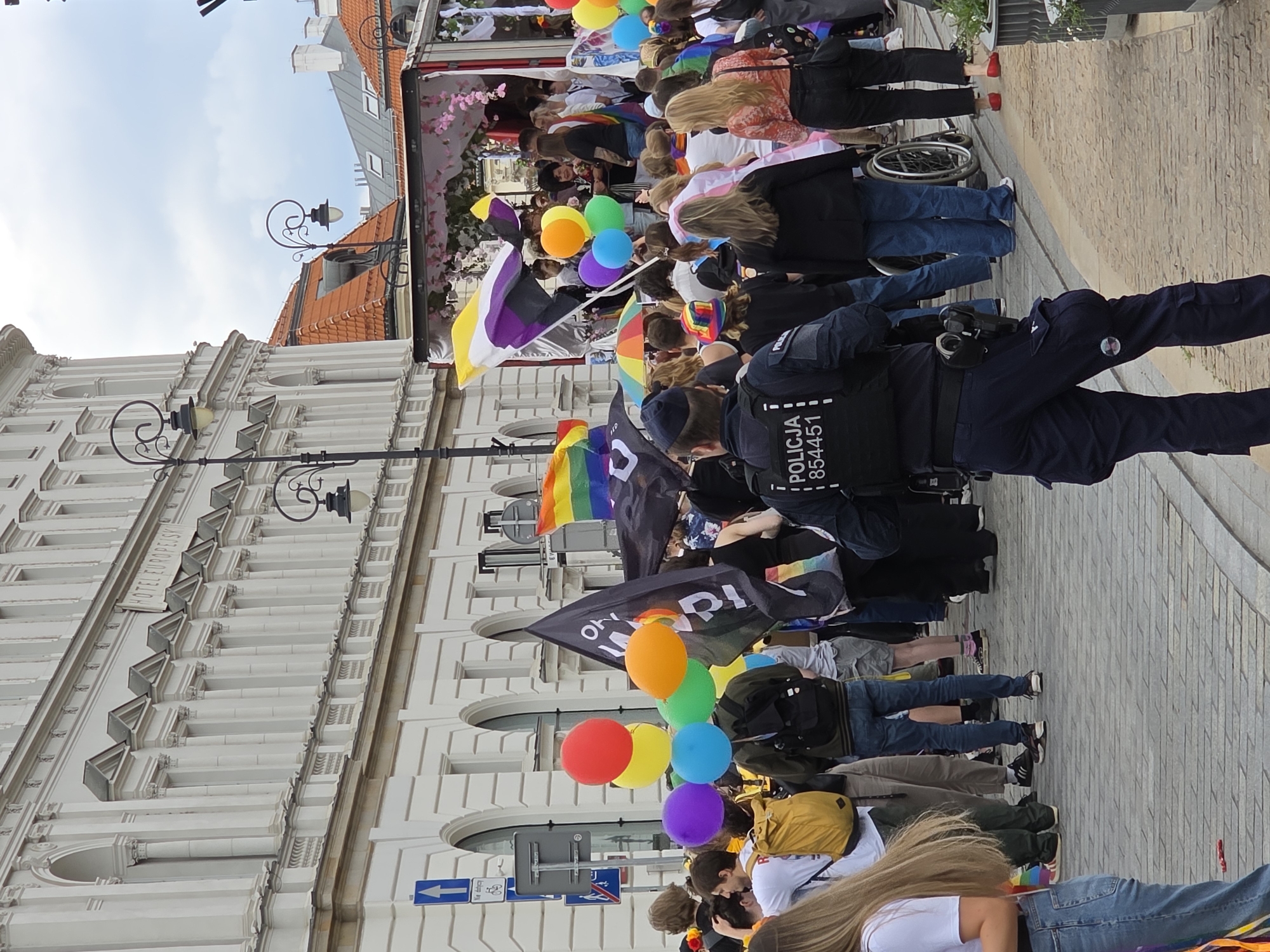 The image depicts a street scene in Warsaw, Poland, likely during a Pride event given the presence of rainbow flags and a general celebratory atmosphere. In the foreground, a police officer wearing a tactical vest with "POLICJA" and the number "854451" printed on it stands facing away from the camera. The officer is equipped with a duty belt holding handcuffs and a firearm. Behind the officer, a diverse crowd of people are gathered.
Several individuals are holding flags, prominently featuring the rainbow flag, a symbol of LGBTQ+ pride, and what appears to be the non-binary pride flag. Balloons in various bright colors, including red, yellow, orange, green, blue, and purple, are also visible, held by some of the participants or tied to objects.
A white building with classical architectural elements, characterized by columns and arched windows, forms the backdrop on the left side of the image. The building has "HOTEL EUROPEJSKI" written across its facade. On the right side, there's a glimpse of a stage or float decorated with floral arrangements. The street is paved with cobblestones. The sky is overcast. The overall scene suggests a public gathering or parade, with a focus on LGBTQ+ visibility and rights.