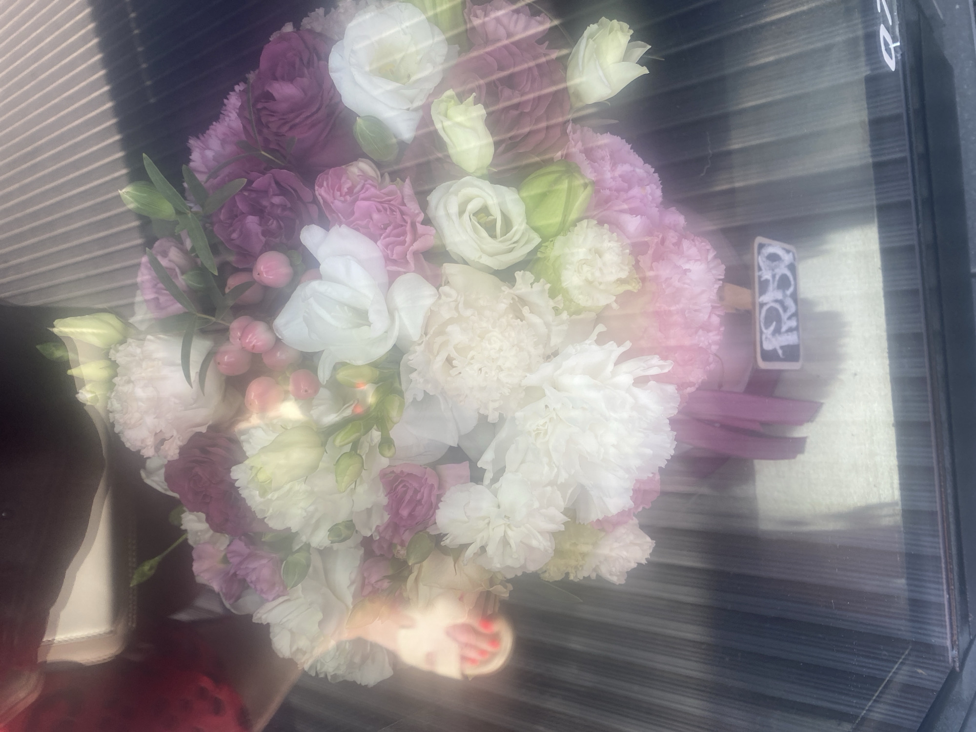 This is a medium shot of a bouquet of flowers in a vase. The flowers are a mix of white, pink, and purple, with some green accents. The bouquet is arranged in a loose, natural style. In the background, there is a person's foot wearing a sandal with red nail polish. The scene appears to be outdoors, possibly on a patio or balcony, given the sunlight and the striped surface beneath the flowers, which resembles blinds or a slatted surface. A small price tag with handwritten text in a white cursive font is attached to the bouquet. The lighting suggests it is daytime. The overall mood is bright and cheerful. The location context provided is Novi Petrivtsi, Ukraine.