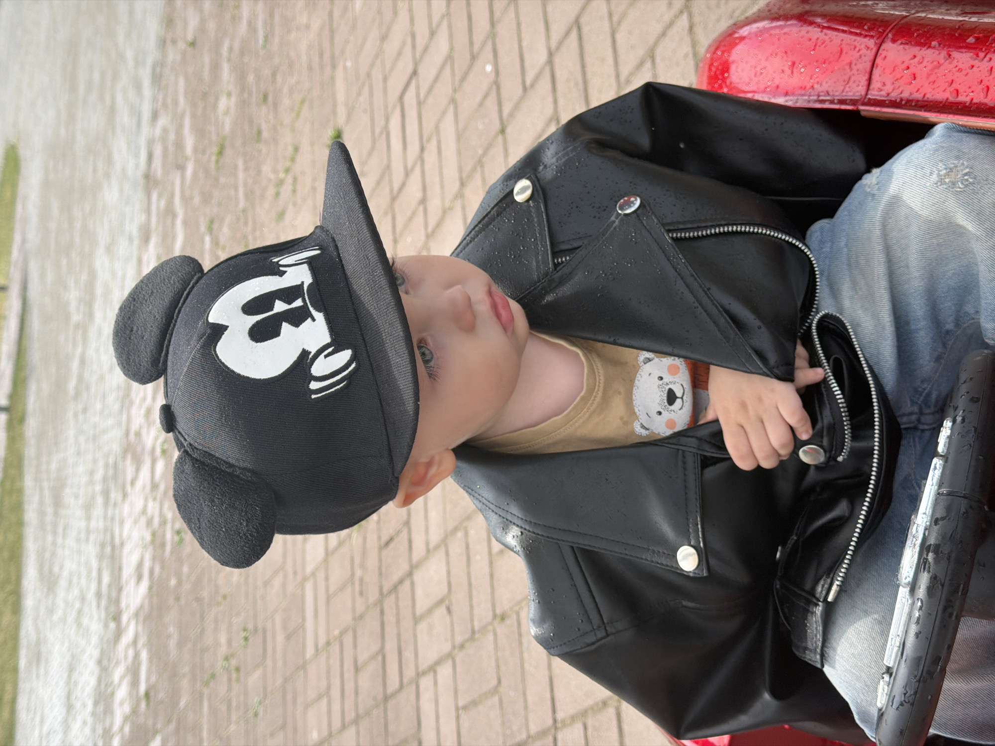 A young child, wearing a black baseball cap with Mickey Mouse ears and a graphic of Mickey Mouse, a tan t-shirt with a bear graphic, a black faux leather jacket with silver snaps and a zipper, and distressed blue jeans, is seated in what appears to be a red toy vehicle. The child is looking to the right, with their left hand partially inside the jacket. The ground is paved with light-colored bricks. The background shows a grassy area and a paved walkway. The scene is outdoors, likely in a park or playground in Chaikovsky, Russia, as indicated by the brick paving and general park-like setting. There are water droplets on the jacket and vehicle, suggesting recent rain.