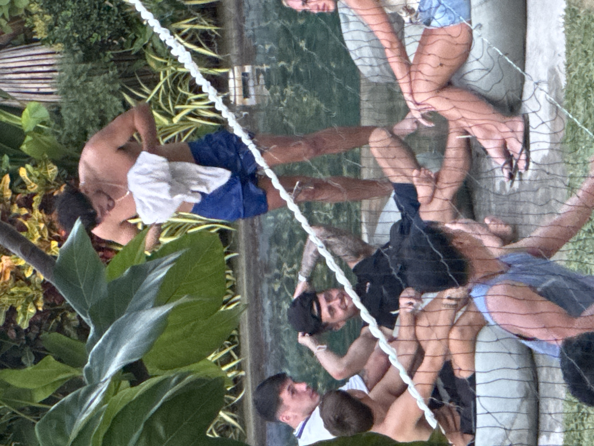 The image, set in General Luna, Philippines, depicts a lively outdoor scene with several young men gathered around a body of water, likely a pool or shallow sea, during daylight hours. Lush green tropical foliage dominates the left foreground, partially framing the activity.
A thick white rope or net, possibly for a game like water volleyball, stretches diagonally across the mid-ground from the upper left to the lower right, separating different groups of people. To the left of this net, a shirtless man wearing blue shorts and with a white cloth draped over his arm is partially visible, his face obscured as he leans forward.
On the right side of the net, multiple individuals are present. A man in a black t-shirt and a black cap stands prominently, looking towards the left and smiling. He has visible tattoos on his arms. Other men are seen around him; one is in a white collared shirt, and another wears a light blue top. Their legs and torsos are visible through the net, some appearing to be partially submerged in the clear water, which shows reflections of the bright sunlight. In the background, a concrete structure and a wooden fence can be seen beside the water. The overall atmosphere appears relaxed and social, indicative of leisure activities in a tropical setting. There is no visible text.