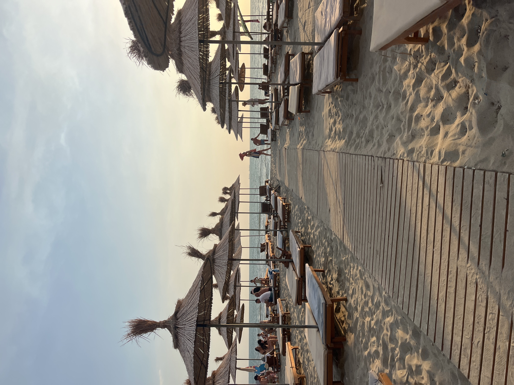 This image depicts a beach scene in Bashkia Durrës, Albania, likely during the late afternoon or early evening, as indicated by the soft, warm lighting. The foreground features a wooden boardwalk leading into a sandy expanse. Rows of empty, cushioned lounge chairs and thatched umbrellas are neatly arranged on the sand, awaiting beachgoers. In the middle ground, a few individuals are visible, some seated on lounge chairs or standing near the water. The sea stretches to the horizon under a pale, partly cloudy sky. The overall atmosphere is serene and peaceful.