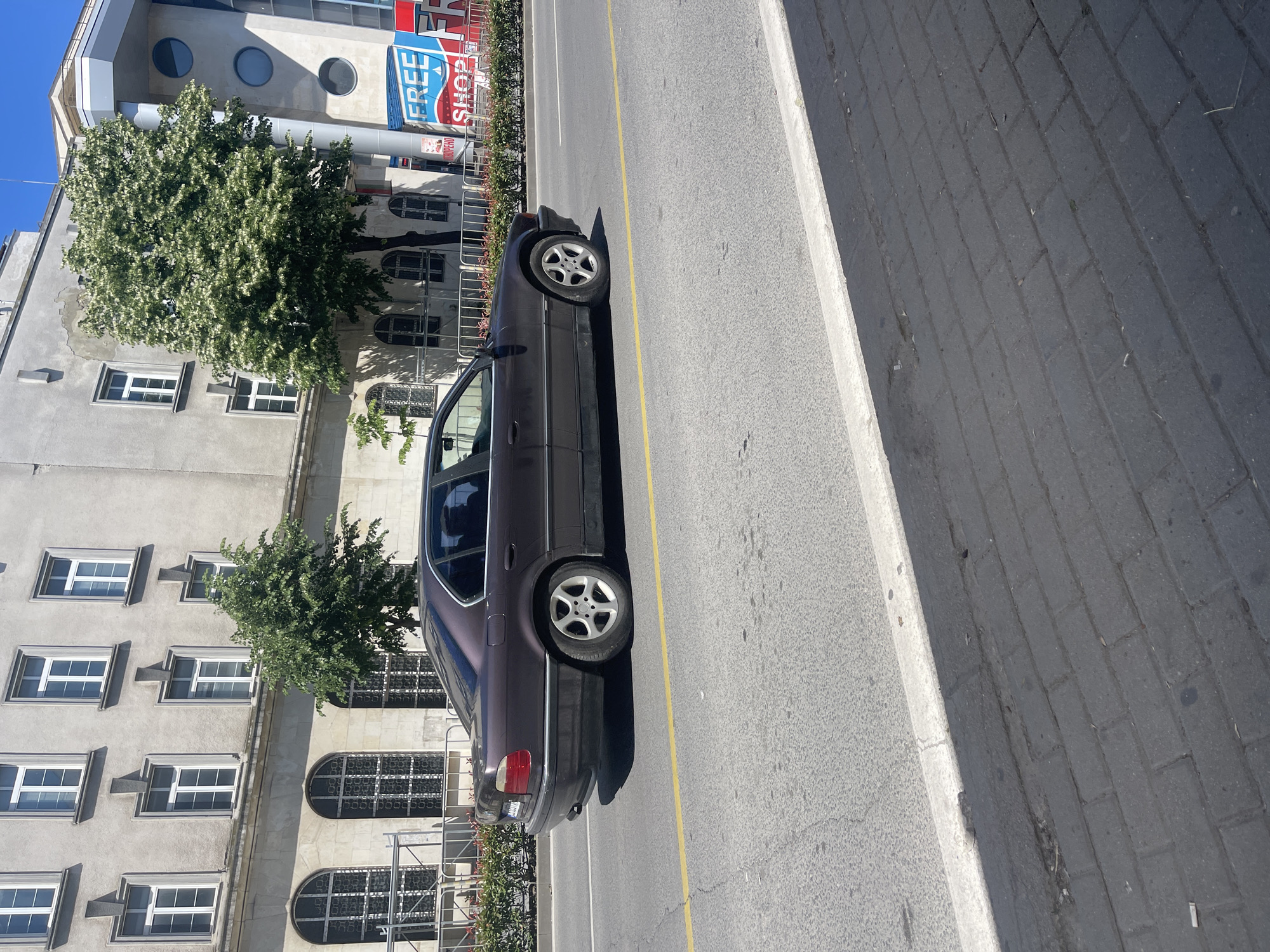 A dark-colored sedan is driving on a street in Varna, Bulgaria. The car is a BMW, likely a 7 Series model from the late 1990s or early 2000s. It is a right-hand drive vehicle, indicating its location in a country that drives on the right. The car is traveling from left to right in the frame. The street appears to be paved with asphalt and has a single yellow lane divider. On the right side of the street, there is a sidewalk made of dark paving stones. Behind the sidewalk and street, there is a building with multiple windows and a modern structure with circular windows and signage that includes the words "FREE SHOP." The building on the left is older, with a light gray facade and numerous rectangular windows. Trees with green foliage are visible between the car and the buildings. The sky is clear and blue, suggesting a sunny day. The scene is captured from a slightly elevated perspective, looking down at the street and car.