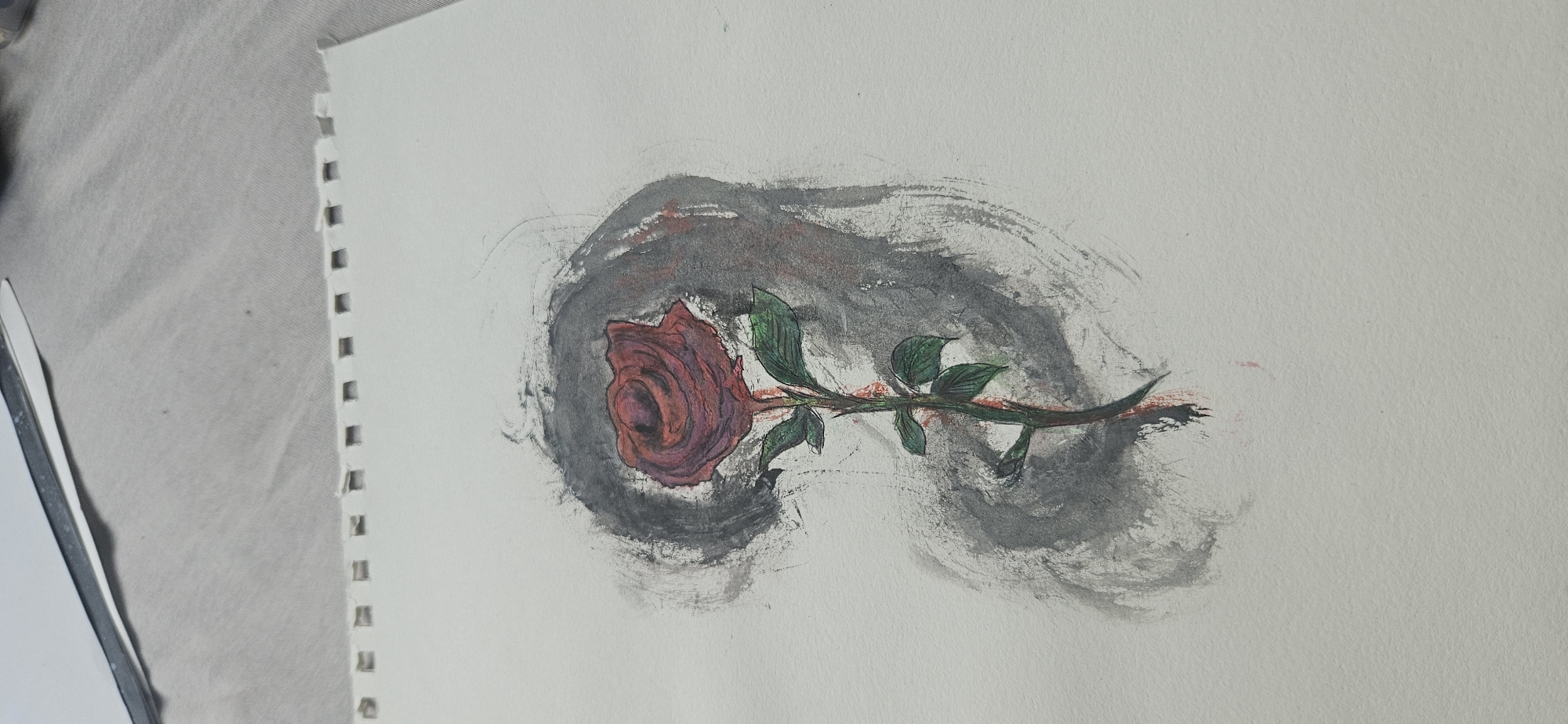 The image shows a drawing of a red rose with a green stem and leaves, lying horizontally on a white piece of paper. The rose is surrounded by a dark, grey, hazy background that is likely painted. The paper appears to be part of a spiral-bound notebook or sketchbook. No people are visible. The location is not discernible from the image; there is no evidence indicating it is in Kostenets, Bulgaria.