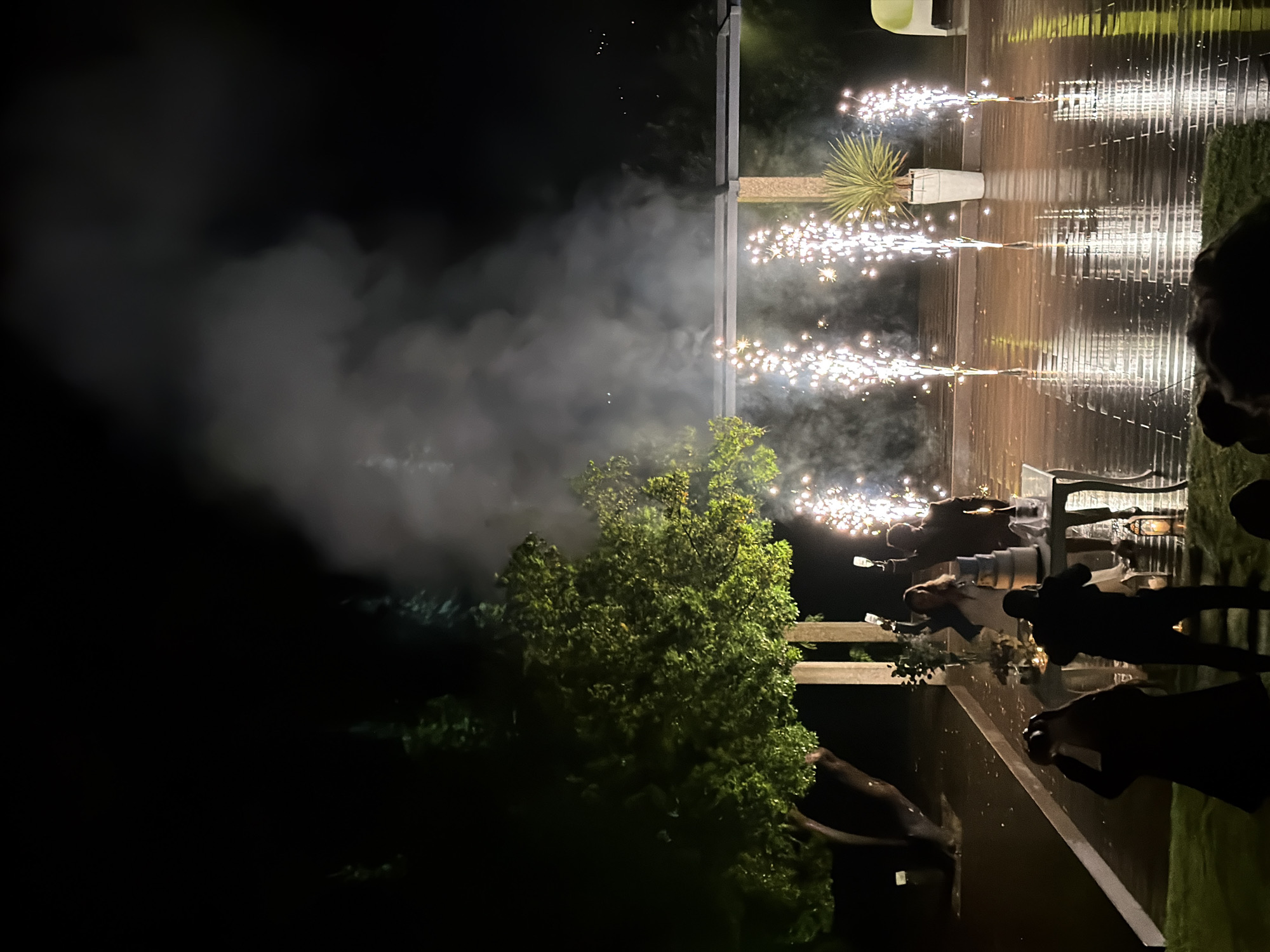 The image depicts a nighttime scene outdoors, likely a celebration, with visible pyrotechnics (sparklers) and several silhouetted figures. A plume of smoke from the fireworks is also present. The setting appears to be a deck or patio area adjacent to a building. One figure is holding a bottle, potentially of champagne. There are plants in pots, and possibly a tree in the foreground. It is difficult to determine if the city of Vila Real, Portugal can be verified as the location based only on visual evidence.