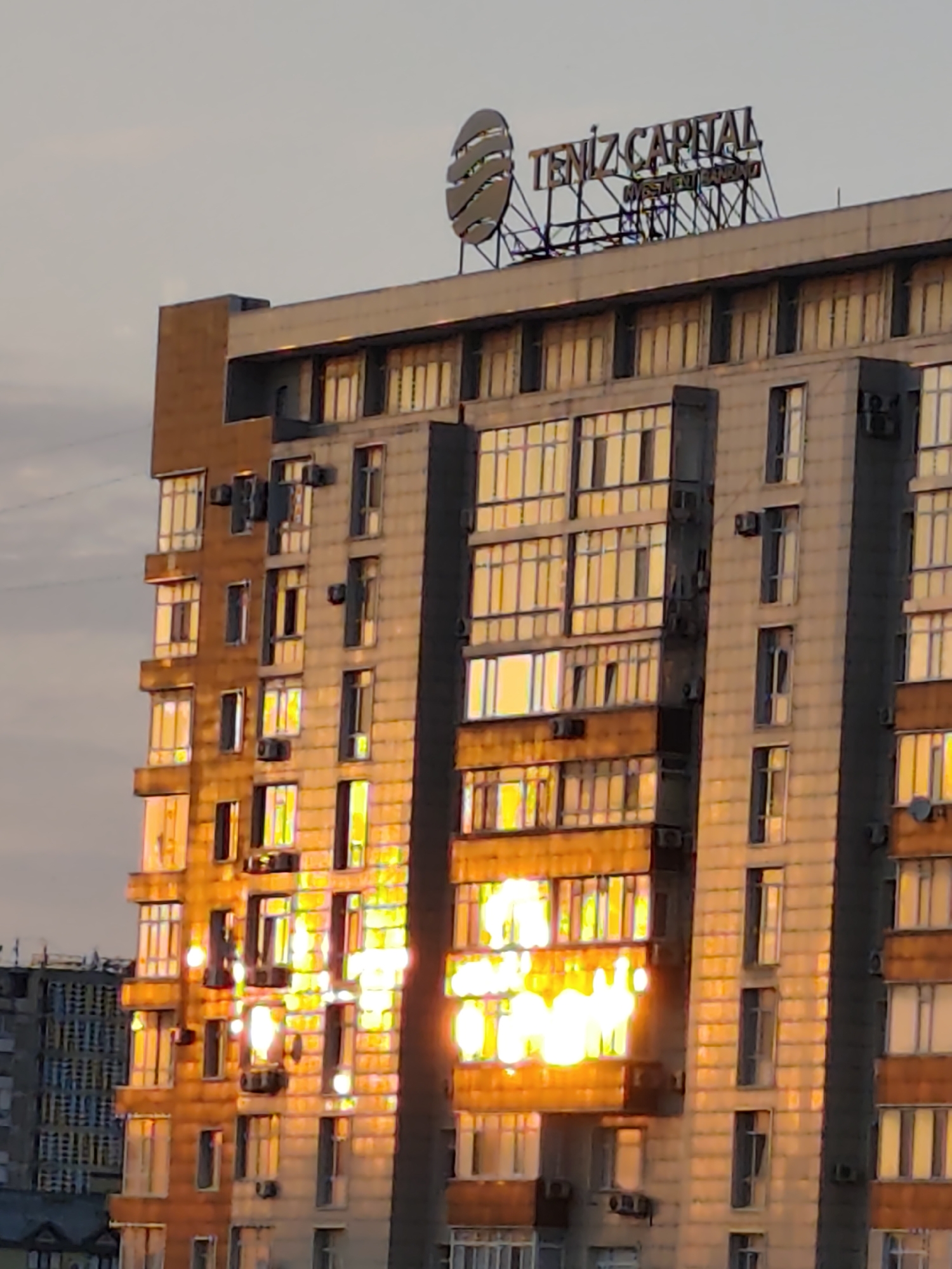 The image shows a close-up view of the upper floors of a modern, multi-story building in Aktau, Kazakhstan. The building features a facade with numerous windows, many of which reflect the bright sunlight, creating a dazzling, golden glow. The windows are arranged in a grid pattern, with some balconies visible. At the top of the building, a large sign with the words "TENIZ CAPITAL" and "INVESTMENT BANKING" is mounted, along with a circular logo. The sky is overcast, suggesting either sunrise or sunset. In the background, parts of other buildings are visible, including a gray building with a more utilitarian design. The overall impression is one of urban architecture bathed in the warm light of the setting or rising sun.