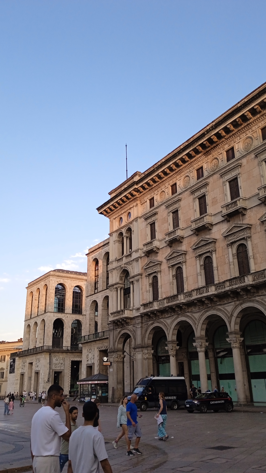 The image shows a view of Piazza del Duomo in Milan, Italy, featuring the Galleria Vittorio Emanuele II and the Palazzo dell'Arco della Pace. The architecture is grand and ornate, with neoclassical elements visible in the buildings. The sky is clear and blue, suggesting it is daytime.
In the foreground, a group of people are walking and standing. Some are dressed casually in t-shirts and shorts, while others are wearing dresses. There is a black van with "CARABINIERI" written on the side, as well as a black Carabinieri vehicle parked in the street. The presence of these vehicles suggests a police presence or a security detail.
The buildings in the background are impressive structures with multiple levels of arches, balconies, and decorative facades. The sunlight casts long shadows, indicating that the sun is low in the sky, possibly late afternoon or early morning. The overall scene is a bustling urban environment with a mix of historical architecture and modern activity.