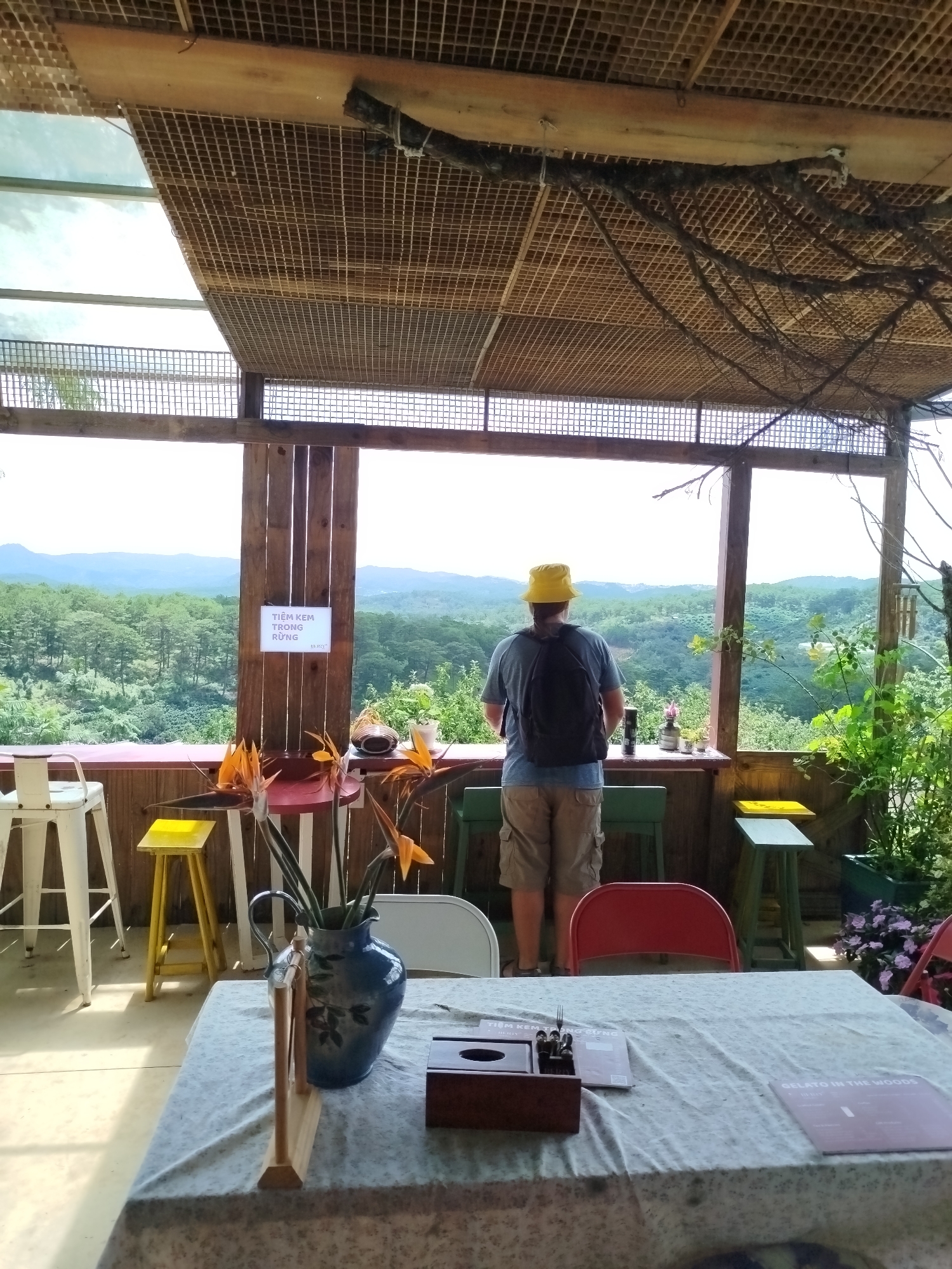 The image depicts an outdoor seating area with a panoramic view of a lush, forested landscape. In the foreground, a table is draped with a patterned tablecloth, and on it sits a blue patterned vase filled with orange and yellow flowers, a wooden napkin holder, and a menu. Behind the table, several chairs are visible, including a red one and two green ones. To the left, a white stool stands near a small yellow stool.
A man wearing a yellow bucket hat, a blue t-shirt, and khaki shorts stands with his back to the camera, looking out at the view. He is wearing a black backpack and holds a silver thermos in his left hand. A sign on a wooden post behind him reads "TIEM KEM TRONG RUNG" in Vietnamese, which translates to "Ice Cream in the Forest." The structure above the seating area is made of wood with a lattice ceiling, and branches hang down. The background features rolling hills covered in dense greenery, indicating a natural or park-like setting, likely in the vicinity of Trường An, Vietnam, known for its scenic beauty. The overall atmosphere is relaxed and natural.
