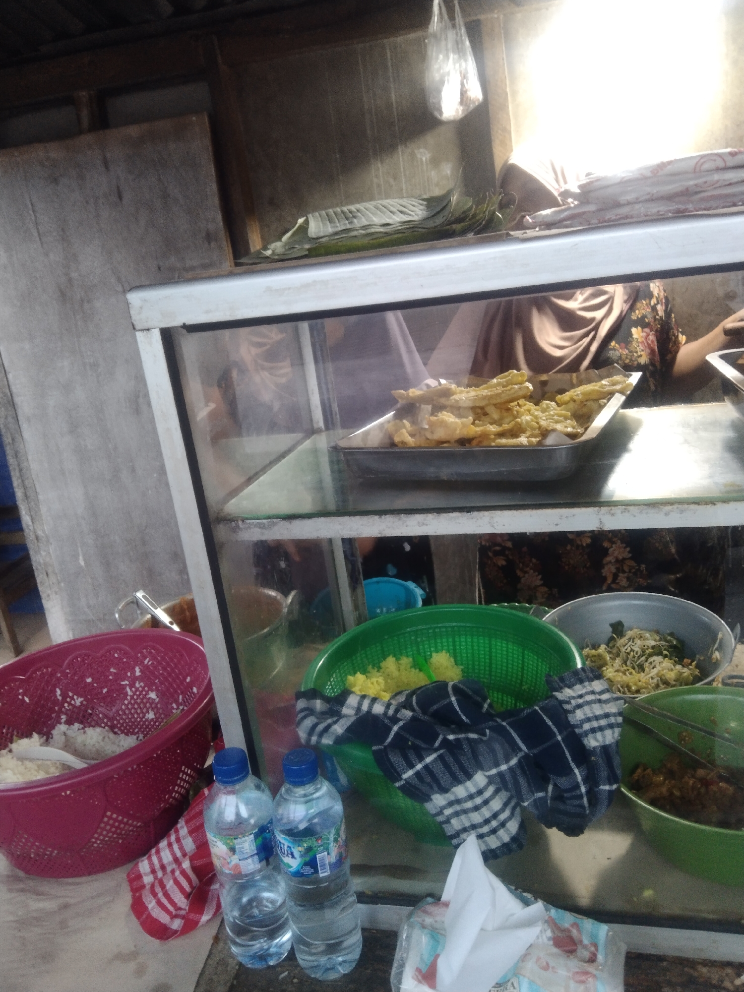 A vendor stands behind a glass display counter filled with food at a street stall in Klepek, Indonesia. In the top shelf of the display, a metal tray holds fried food items, possibly fritters or snacks. Below, there are several bowls containing prepared food, including what appears to be rice in a pink woven basket, a green basket with yellow food, and a grey bowl with a mixture of vegetables and noodles. A dark blue and white checkered cloth is draped over the green basket. To the right of the counter, two plastic water bottles sit on a surface, next to a red and white checkered cloth. The stall appears to be set up outdoors, with a rustic wooden structure forming the background. Sunlight streams in from the right, creating bright highlights on the display counter.