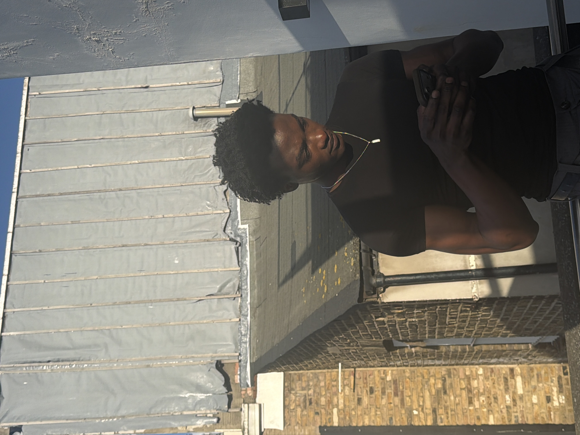A man stands on a rooftop in London, UK, talking on a cell phone. The roof is covered in gray, tarpaulin-like material and the surrounding buildings are brick. The man is wearing a black t-shirt and dark-colored pants. He has dark hair and a gold necklace. He is holding the phone to his ear with his left hand. The overall setting is urban and industrial.