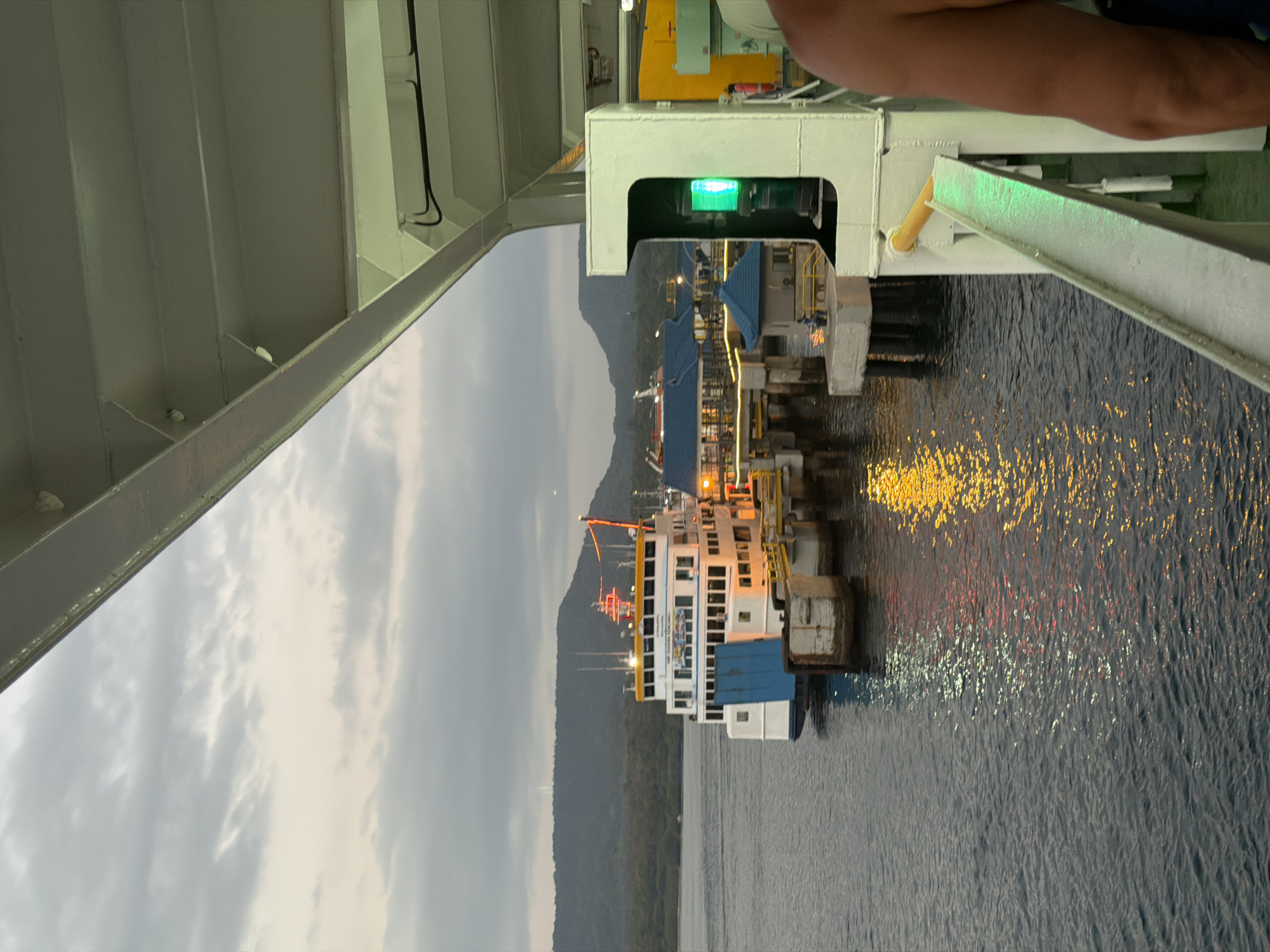 An outdoor, medium-shot, eye-level, vertical, full-color, natural light photograph captures a ferry docked at a pier in the city of Melaya, Indonesia. The ferry, predominantly white with blue accents, is positioned to the left of the frame, its lights reflecting on the dark water. Several piers and structures, including a small blue-roofed building, are visible extending from the shoreline. A mountainous landscape with lush vegetation forms the backdrop under a cloudy, twilight sky. The foreground is occupied by the perspective from another vessel, with a white railing and a green navigation light prominently featured on the right. The scene suggests a port or harbor environment during the evening hours.