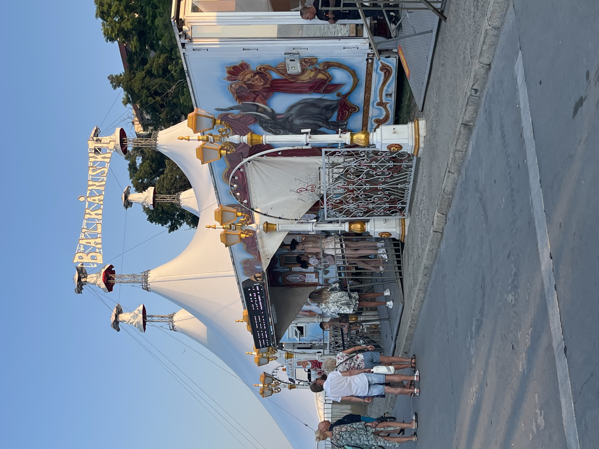 A group of people are gathered outside a circus tent in Nesebar, Bulgaria. The tent is white with prominent golden decorations, including ornate lamp posts and a sign that reads "Balkanski". A large truck with colorful murals of an elephant and a crowned figure is parked next to the tent. Several individuals are standing in line or mingling near the entrance. The scene is set against a clear blue sky and a backdrop of trees and buildings on a hillside.