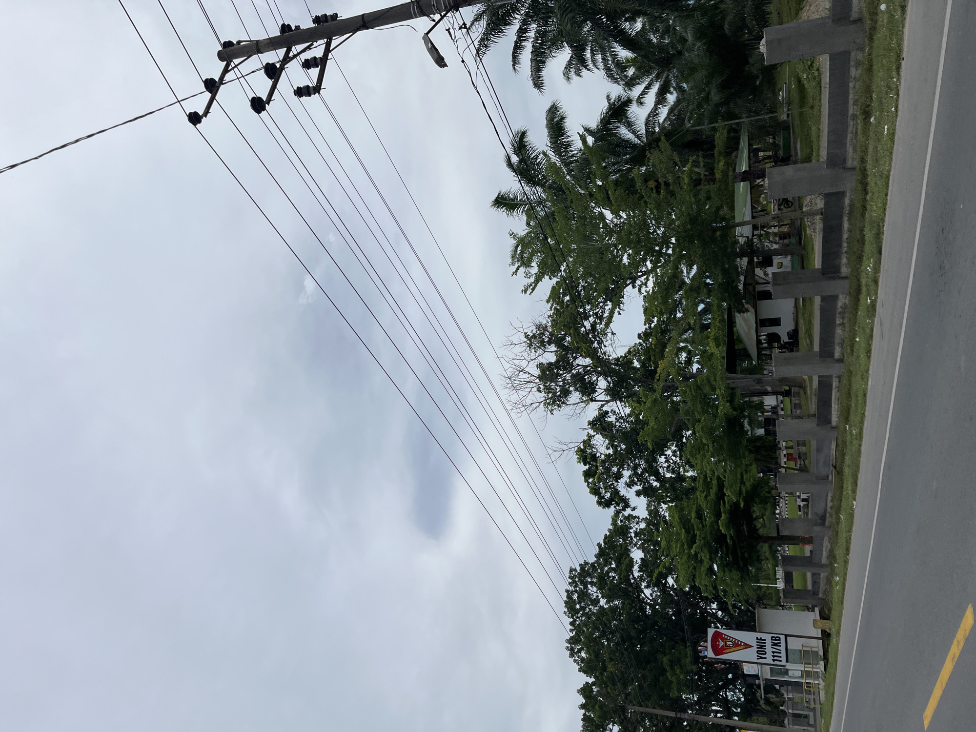 This image depicts a street scene in Simpangempat, Indonesia, on a cloudy day. A paved road with a yellow line runs along the bottom of the frame. To the left of the road, there is a sign with the text "YONIF 111/KB" and a logo. Beyond the sign, a low concrete wall runs parallel to the road, with several concrete posts. Lush green trees and palm trees are visible behind the wall, some obscuring buildings. A utility pole with multiple wires stretching across the frame is prominent on the right side, with a street lamp attached to it. The sky is overcast and gray, suggesting a potentially humid or rainy climate. No people are visible in the image.