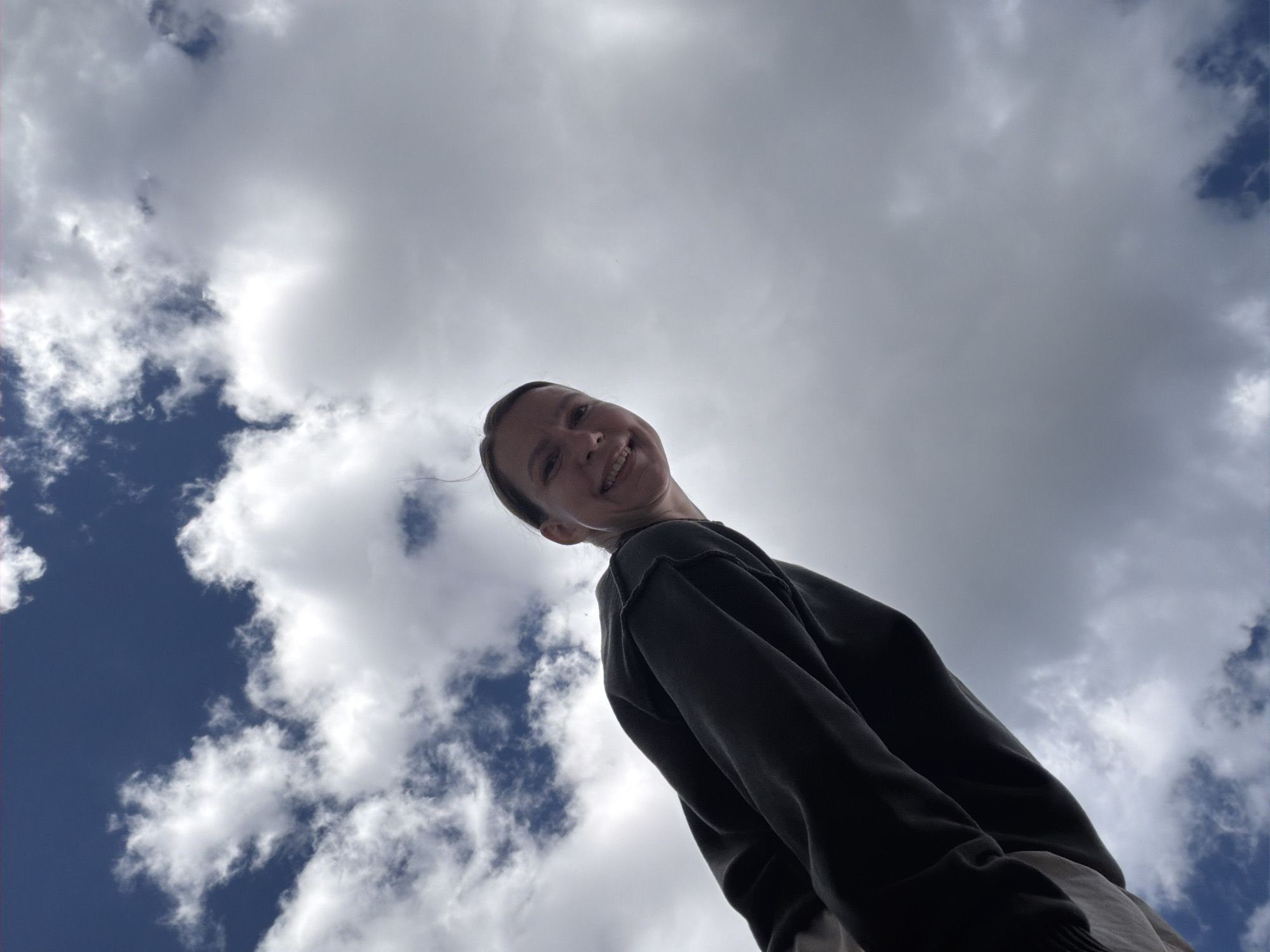A low-angle medium shot depicts a smiling person against a sky filled with clouds. The person is wearing a dark-colored long-sleeved shirt. The sky alternates between patches of blue and large, puffy white clouds. There's no information available to deduce the location as Chaikovsky, Russia.
