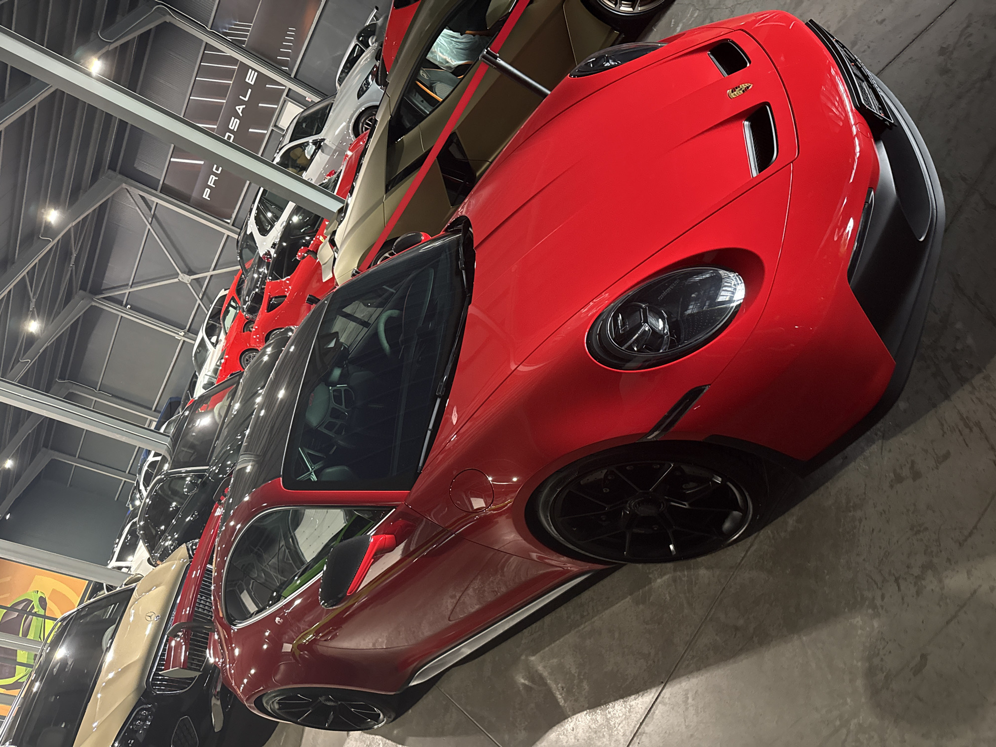 The media file shows a collection of cars, predominantly red, parked in a well-lit indoor space. The cars are lined up, suggesting a showroom or a storage facility. The main subject is a bright red sports car in the foreground, angled towards the viewer. Its sleek design, black rims, and prominent headlights are clearly visible. Behind it, other cars can be seen, including a white and a beige car, also sports car models, and darker colored vehicles further down the line. The setting appears to be a modern industrial building with exposed metal beams and ceiling lights. A sign with white lettering on a dark background is partially visible in the upper left, reading "PRO SALE". The overall impression is one of a high-end automotive collection, possibly a dealership or a private enthusiast's garage. The lighting is bright and even, indicating artificial illumination, and the floor is a smooth, grey concrete surface, reflecting some of the light. There are no people visible in the frame. The angle of the shot is from a slightly elevated viewpoint looking down at the cars. The time of day is not discernible due to the indoor setting and artificial lighting. The location is Plovdiv, Bulgaria.