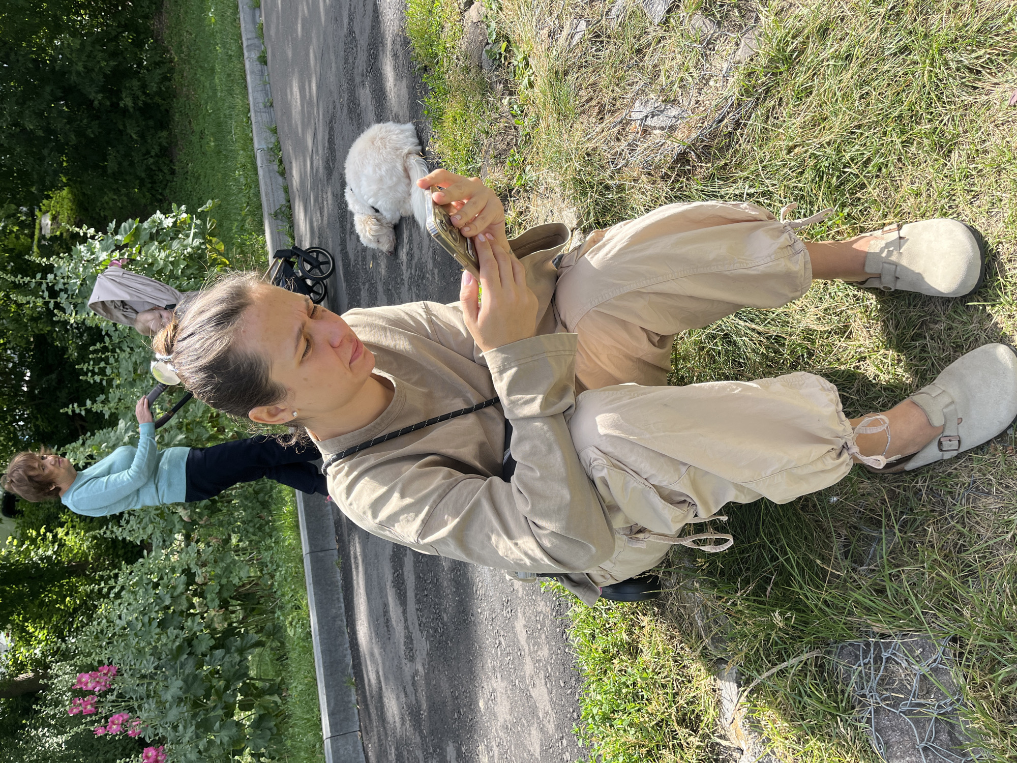 A woman in tan, baggy cargo pants and a long-sleeved tan shirt sits on a patch of grass, looking at her phone. She wears light tan clogs. Her hair is pulled back into a bun. Behind her, a white, fluffy dog sleeps curled up on the pavement. Further back, a woman in a light blue sweater and dark pants pushes a stroller with a baby inside. The stroller is next to a bush with pink flowers. The scene appears to be a park or a residential area in Moscow, Russia, with trees, grass, and pavement visible. The lighting suggests it is daytime.