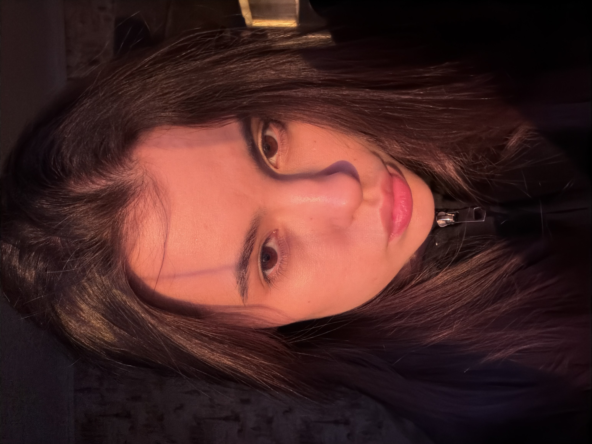 This close-up portrait features a young woman with long, dark brown hair, looking directly at the viewer. Her head is tilted slightly, and her face is illuminated by a warm, reddish-orange light source, casting a soft glow on her skin and hair. Her dark brown eyes are wide open, and her lips are slightly parted, revealing a natural pink color. Her prominent nose is also clearly visible. She wears a dark garment, possibly a jacket, indicated by a metallic zipper pull near her neck. The background is mostly dark and out of focus, suggesting an indoor setting with low light conditions or an intentional blurring effect. There are no specific location cues visible within the image itself, nor is there any discernible weather. Her expression is neutral but direct and observant. No visible text is present. The overall atmosphere is intimate and softly lit.