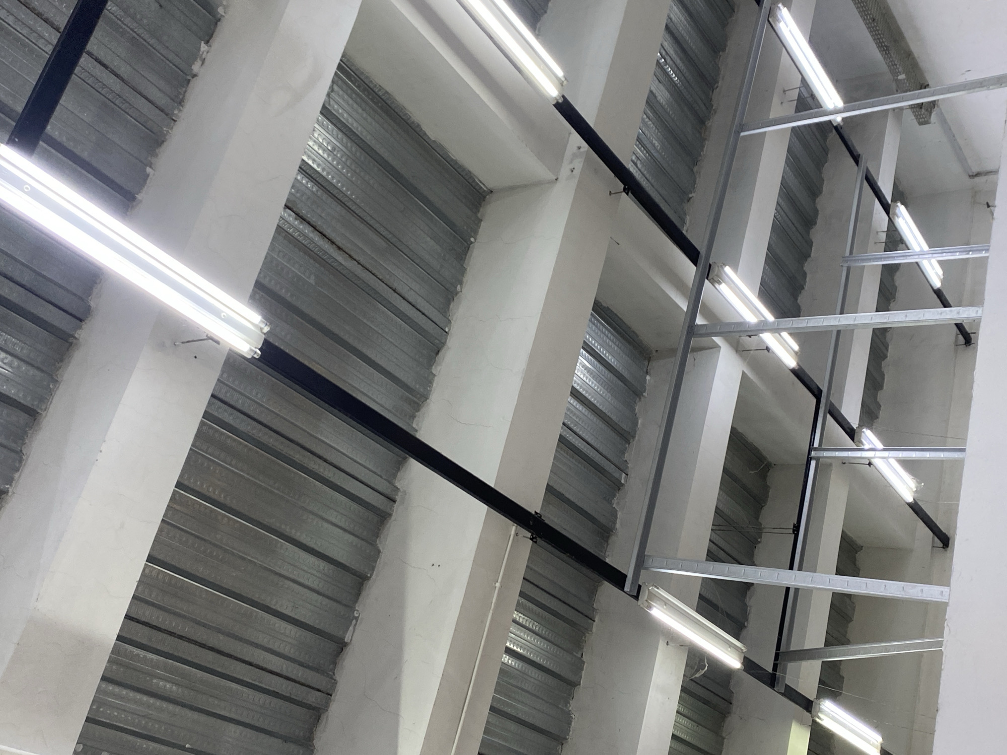 The image displays the interior of a building under construction or renovation, possibly a warehouse or industrial space, located in Depok, Indonesia. The ceiling consists of corrugated metal sheeting supported by white beams and black metal struts. Fluorescent light fixtures are installed throughout the ceiling area. Some sections show exposed metal framing, indicating the ongoing construction. The lighting suggests an indoor environment. There are no people visible in the image.