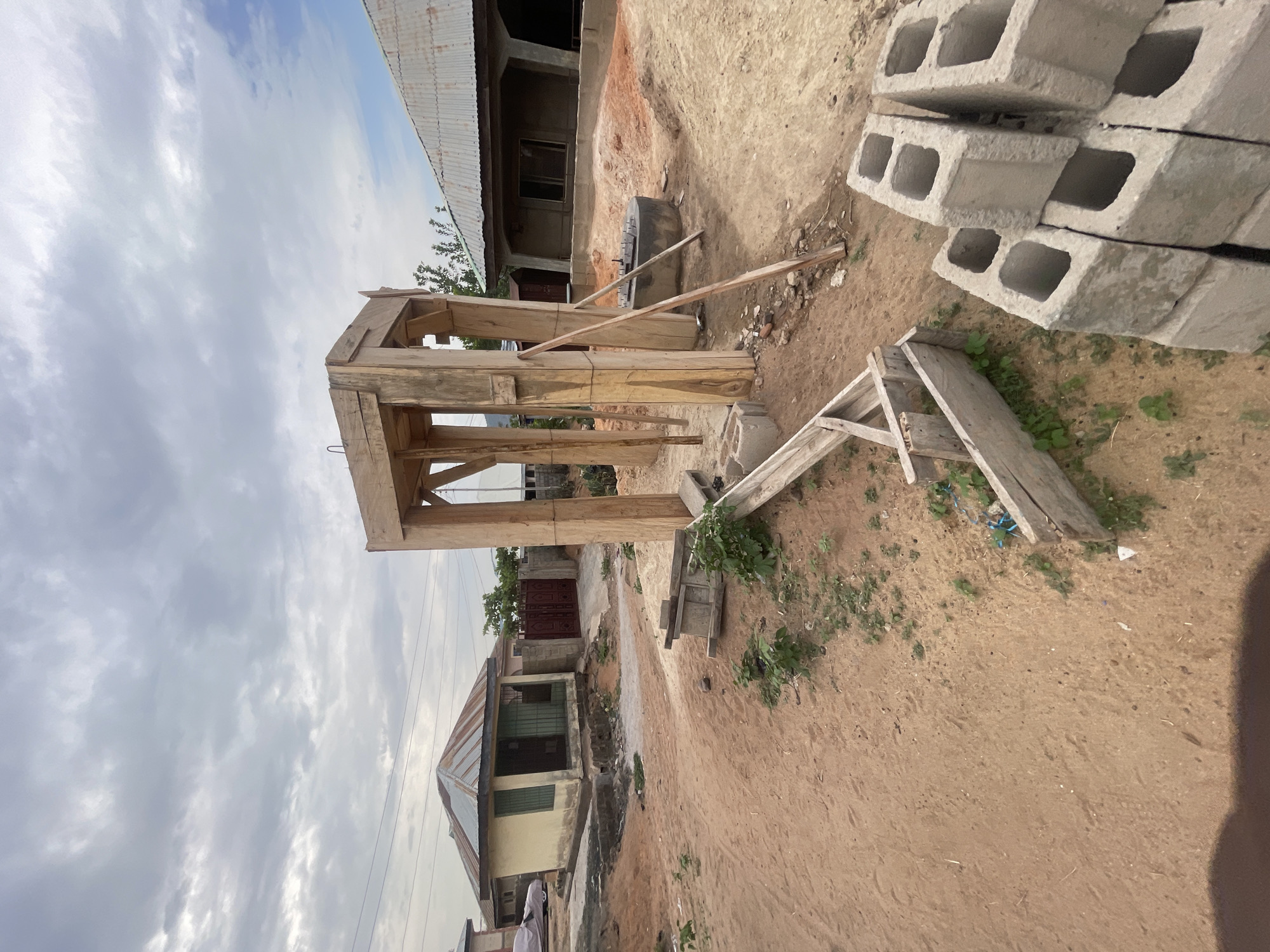 This outdoor image, captured in Lokoja, Nigeria, depicts a scene of ongoing construction or building activity under a cloudy daytime sky. No people are visible in the frame.
The central subject is a robust wooden structure, likely formwork or a temporary support system for concrete, constructed from rough timber beams. To the right of this structure, a neatly stacked pile of gray concrete hollow blocks (cinder blocks) rests on the reddish-brown dirt ground. Adjacent to the blocks, a smaller, roughly built wooden frame, possibly a makeshift ladder or support, lies on the ground. Near the base of the main wooden structure, a dark, cylindrical object, resembling a large tire or a foundation form, is present. Several loose wooden planks are scattered across the uneven, earthy terrain, which also features sparse patches of green vegetation.
In the background, on the left, a single-story building with a corrugated metal roof, light-colored walls, and green-framed windows is partially visible. Further back, a dark, ornate metal gate indicates another property entrance. On the right side of the background, an unfinished building with a concrete facade and another corrugated metal roof suggests additional construction. The sky above is largely overcast with white and gray clouds, indicating a cloudy or partly cloudy day with ample ambient light. There is no visible text in the image. The overall setting suggests a developing or semi-urban area.