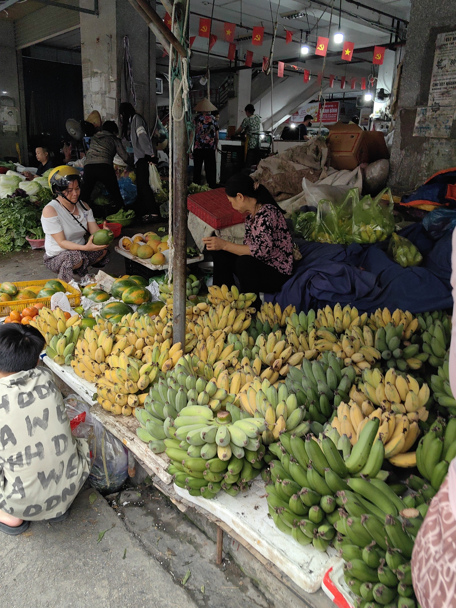 The image depicts a vibrant, busy scene at a market, likely in Ha Long Ward, Vietnam. Predominantly featured are large quantities of bananas in varying stages of ripeness, from green to yellow, arranged on a white-topped display table. Other fruits like mangoes and papayas are scattered around the bananas. Two women appear to be vendors, one seated, wearing a floral top, and the other crouching, wearing a white shirt and a helmet. A child in a patterned jacket is also present near the fruit display. In the background, several people are moving about, some of whom are interacting with the merchandise. The market area is somewhat enclosed, with concrete pillars and various goods stored in bags and containers. Red flags with a yellow star are strung across the upper part of the scene, and a sign that reads "ẢNH BÌNH" can be seen in the background. Another sign with the text "CHO VAY NHANH" is also visible in the upper right corner of the image. The overall atmosphere is bustling, with a focus on fresh produce being sold at a local market.