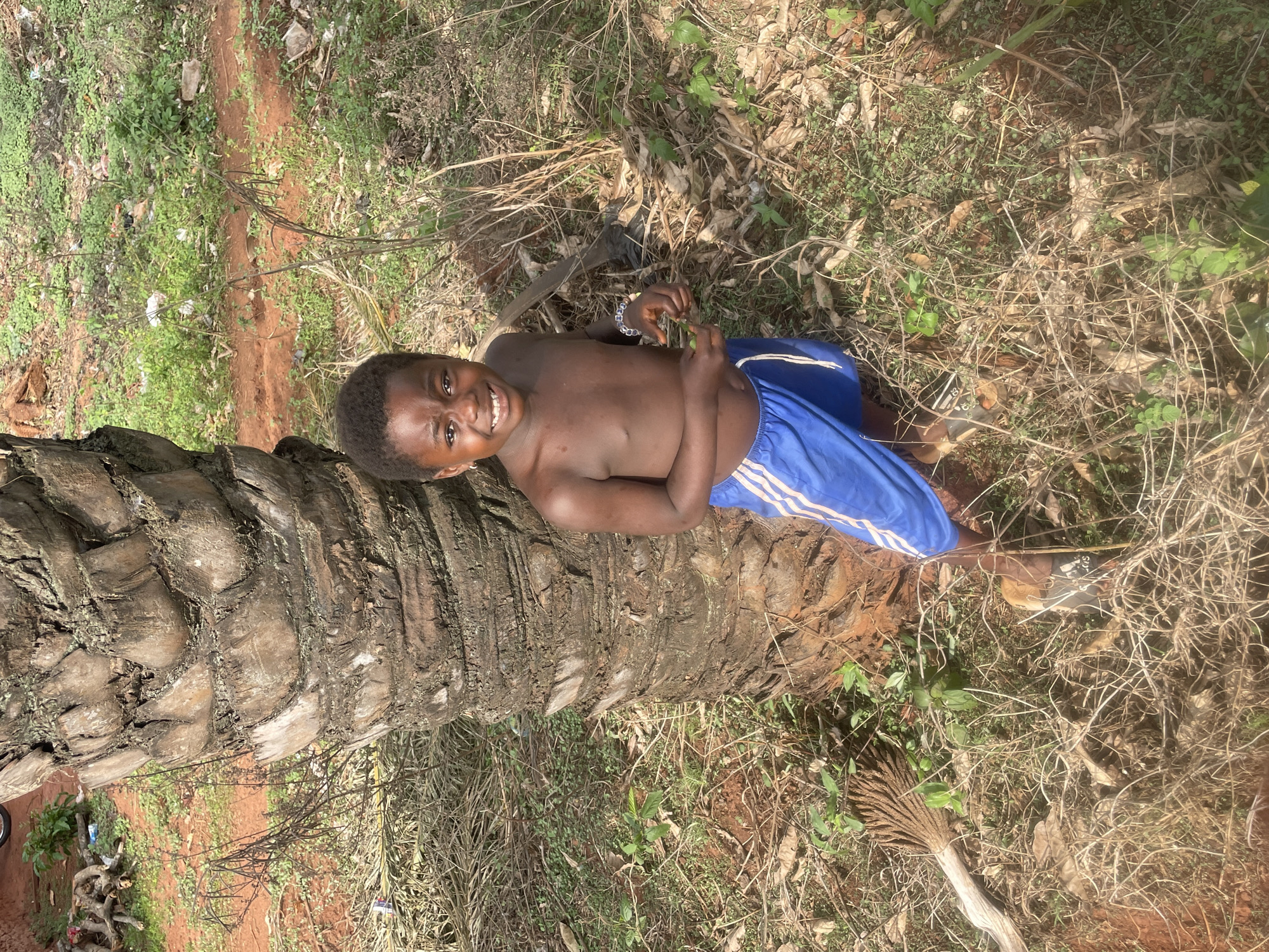 A young, shirtless boy is lying horizontally on the trunk of a felled palm tree in Ogbe, Nigeria. He is smiling and wearing blue shorts with white stripes down the side. The trunk of the palm tree runs vertically and is surrounded by dry grass, small green plants, and scattered leaves. A pair of shoes and a small palm flower are visible in the lower right corner of the frame. The location appears to be an area with natural vegetation and red soil.