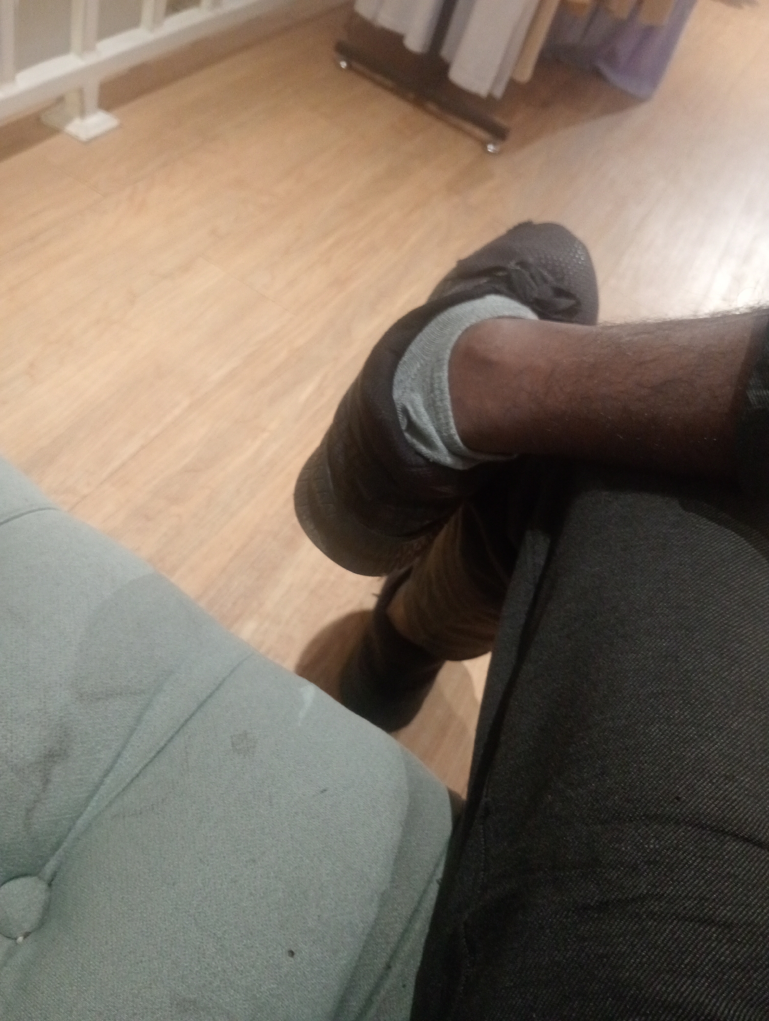 A close-up, eye-level shot shows a person's legs and feet, with the left leg crossed over the right. The person is wearing a dark gray sock and a dark-colored sneaker on the left foot, and dark pants on the right leg. The person appears to be sitting on a light blue tufted surface, possibly a chair or sofa. The floor is made of light-colored wood planks. In the background, a white railing is visible, and beyond that, some clothing items on hangers. The scene suggests an indoor setting, possibly a retail store or a waiting area. The specific location in Larangan, Indonesia is not identifiable from the visual information provided in the image.