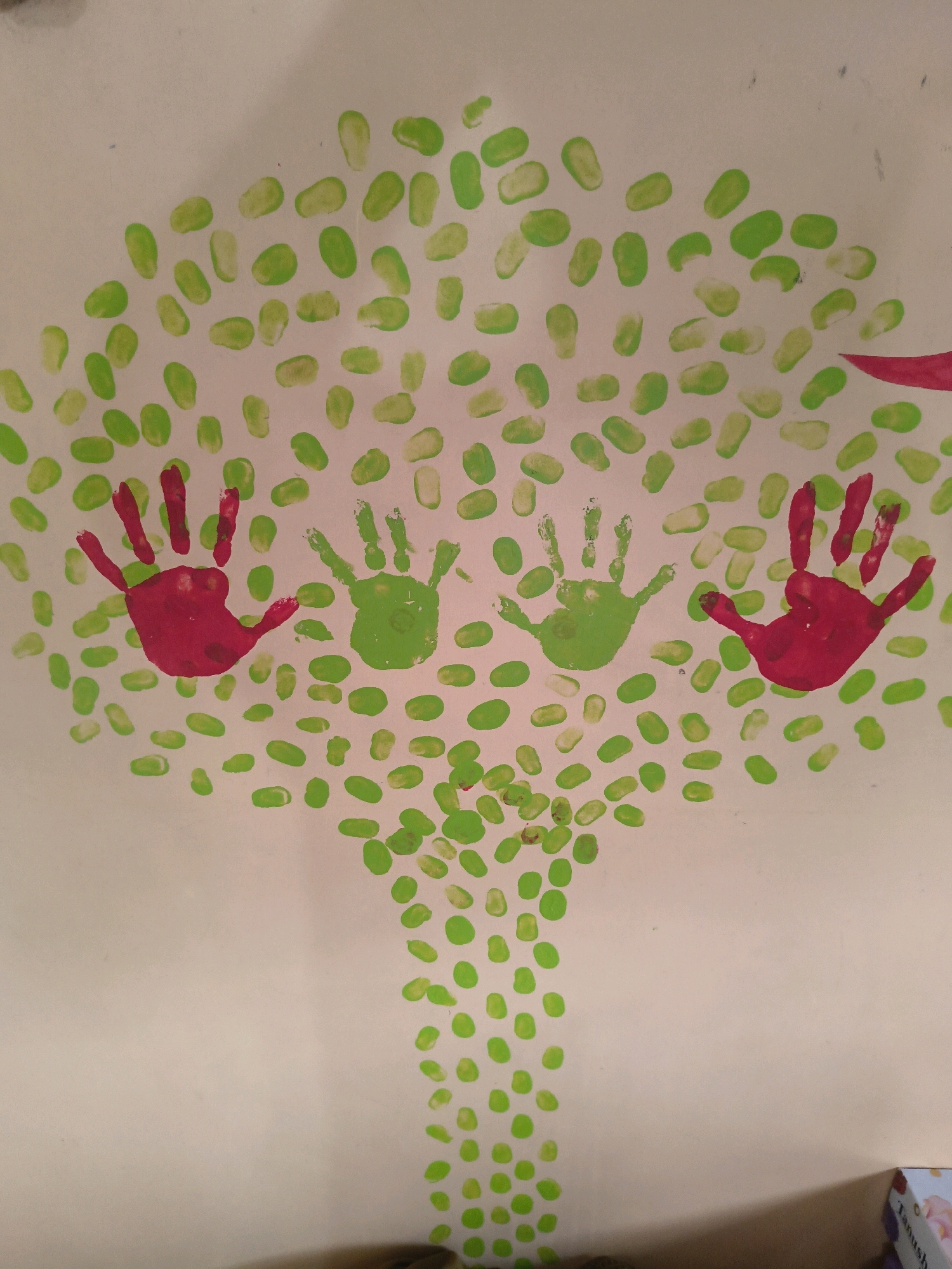 This is a close-up, medium-angle, eye-level shot of a piece of artwork created with paint. The artwork depicts a stylized tree with a trunk made of numerous small green dots, resembling fingerprints or daubs of paint. The top of the tree, the canopy, is also formed by a dense scattering of these small green dots, creating a leafy effect. Prominently featured within the canopy are two handprints. The left handprint is a vibrant red, and the right handprint is a bright green. Both handprints are palm-down, with fingers extended upwards. The background is a plain, light-colored surface, likely paper or canvas. The lighting is even, suggesting an indoor environment. The image is centrally focused on the handprints and the surrounding green dots. There is no indication of the city of Boisar, India, or any specific location within it. The artwork appears to be a child's craft project.