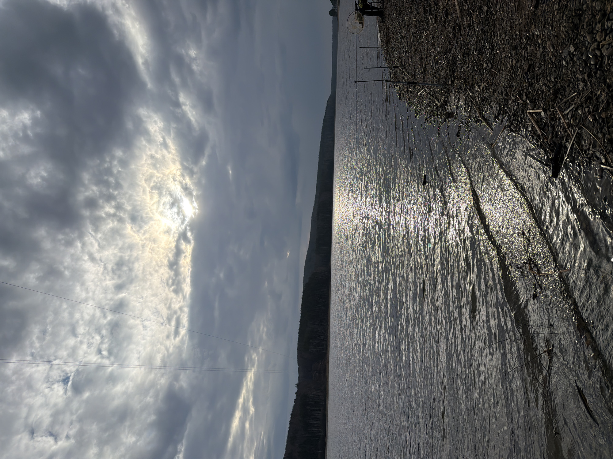 The image captures a tranquil outdoor scene featuring a large body of water, a distant forested bank, and a cloudy sky with strong sunlight. The perspective is from a low vantage point on a rocky or pebbly shoreline, looking across the water towards the horizon.
The sky, occupying the upper portion of the frame, is largely covered by grey and white clouds. A prominent bright patch of sunlight breaks through the clouds on the left side, casting a strong glare across the water below. Two thin, dark utility lines stretch horizontally across the upper-left quadrant of the sky.
In the middle distance, a dark, forested hillside or mountain range runs horizontally, outlining the far bank of the expansive body of water. The trees appear dense and dark, possibly silhouetted against the brighter sky and water.
The lower half of the image is dominated by the water's surface, which intensely reflects the sunlight, creating a shimmering, almost glittering effect across its expanse. Ripples are visible across the water. The immediate foreground on the right features a dark, rocky or pebbly bank, strewn with some natural debris like twigs. Several fishing rods are set up vertically along this bank, firmly planted in the ground, and a circular fishing net or basket hangs from a nearby stand, indicating fishing activity. A small, dark object, possibly a bird, floats on the water's surface near the right bank, and a water strider insect can be seen on the water closer to the shore.
The time of day appears to be daylight, likely morning or afternoon, characterized by partial cloud cover and periods of bright sun. The scene evokes a peaceful and natural atmosphere. No people are visible, and there is no discernible text in the image. The landscape is consistent with natural settings found in Chaikovsky, Russia.
