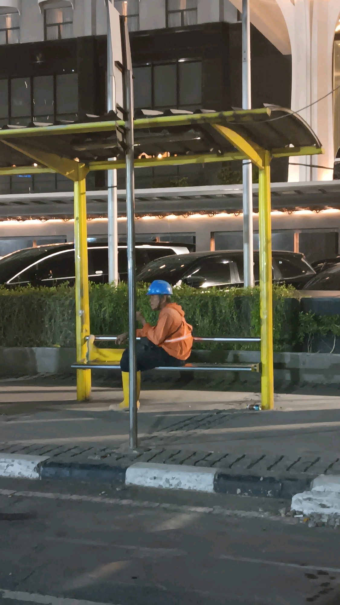 A person wearing a blue hard hat, an orange hooded jacket, black pants, and yellow rubber boots sits on a yellow bench at a bus stop. The person appears to be holding a phone in their right hand. The bus stop has a yellow frame and a dark roof. In the background, there are several black cars parked behind a hedge. The scene is illuminated by artificial lights, suggesting it is nighttime or dusk. The bus stop is located on the side of a street with a concrete curb and asphalt pavement. There is some debris on the ground near the bus stop. The architecture of a building is visible in the upper portion of the image. This scene is consistent with a street-level view in Central Jakarta, Indonesia, likely in an urban or commercial area.