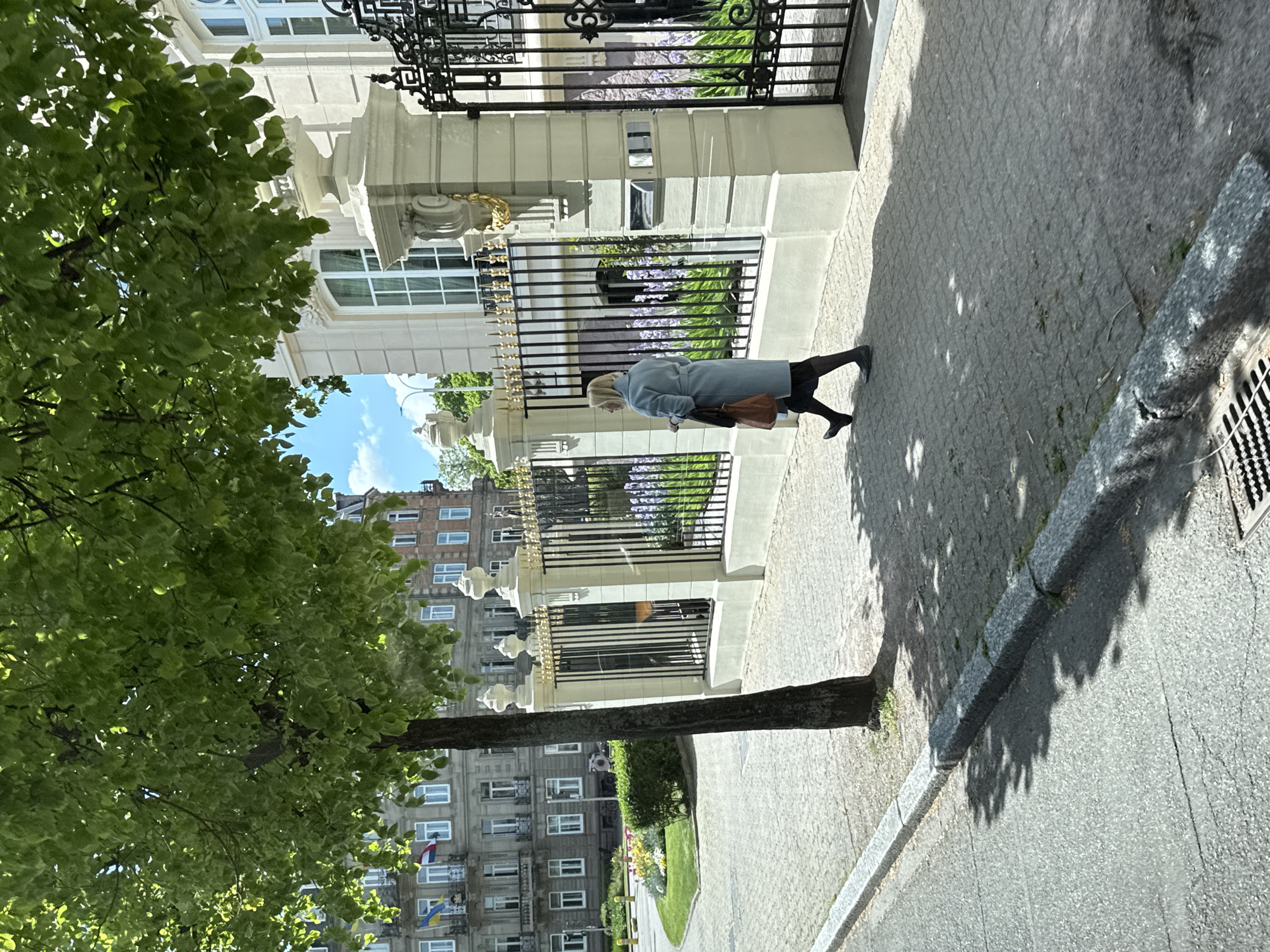 This media file captures a vibrant street scene in what is identified as Strasbourg, France, on a bright, sunny day. The image is oriented sideways, but when viewed upright, it shows an urban landscape with architectural details and a solitary pedestrian.
In the foreground, a woman with blonde hair, dressed in a light blue coat, black skirt, and black boots, walks along a paved sidewalk with a cobblestone pattern. She carries a brown handbag over her left arm and appears to be walking away from the viewer, looking slightly down or to the side.
To her left, a grand, light-colored building with large windows and an elaborate black and gold wrought-iron fence stands prominently. Behind the fence, green foliage and purple flowering plants are visible, suggesting a well-maintained garden or courtyard. Further to the left, a large, leafy green tree partially frames the scene, casting dappled shadows on the sidewalk.
Across the street, in the background, a multi-story residential or commercial building with numerous windows is visible. Notably, two flags are displayed from this building; one clearly shows the blue and yellow stripes of the Ukrainian flag, and another, partially obscured, has red, white, and blue stripes. The street itself, made of asphalt, includes a stone curb and a metal drain grate on the far right. The strong shadows cast by the trees and buildings indicate a clear sky and bright sunshine, likely during midday or early afternoon. No visible text is discernible in the image.
