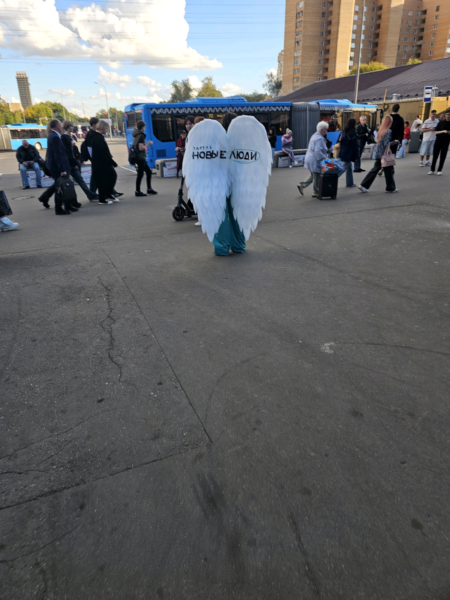 The image shows a street scene in Khimki, Russia, with a person wearing large white angel wings as the central focus. The wings have text on them that appears to read "ЗАРТЕХ НОВЫЕ ЛЮДИ" in black. The person is standing on asphalt, wearing a teal-colored garment that extends to the ground. In the background, several people are walking, some with luggage, suggesting a public transport hub or busy pedestrian area. Blue buses are visible, one with "70" indicated on its side, likely denoting a bus route number. Tall, brick buildings are present in the background, typical of urban architecture. The sky is partly cloudy with a bright blue hue, indicating daytime. The overall scene captures a moment of public activity with an unusual costumed individual.