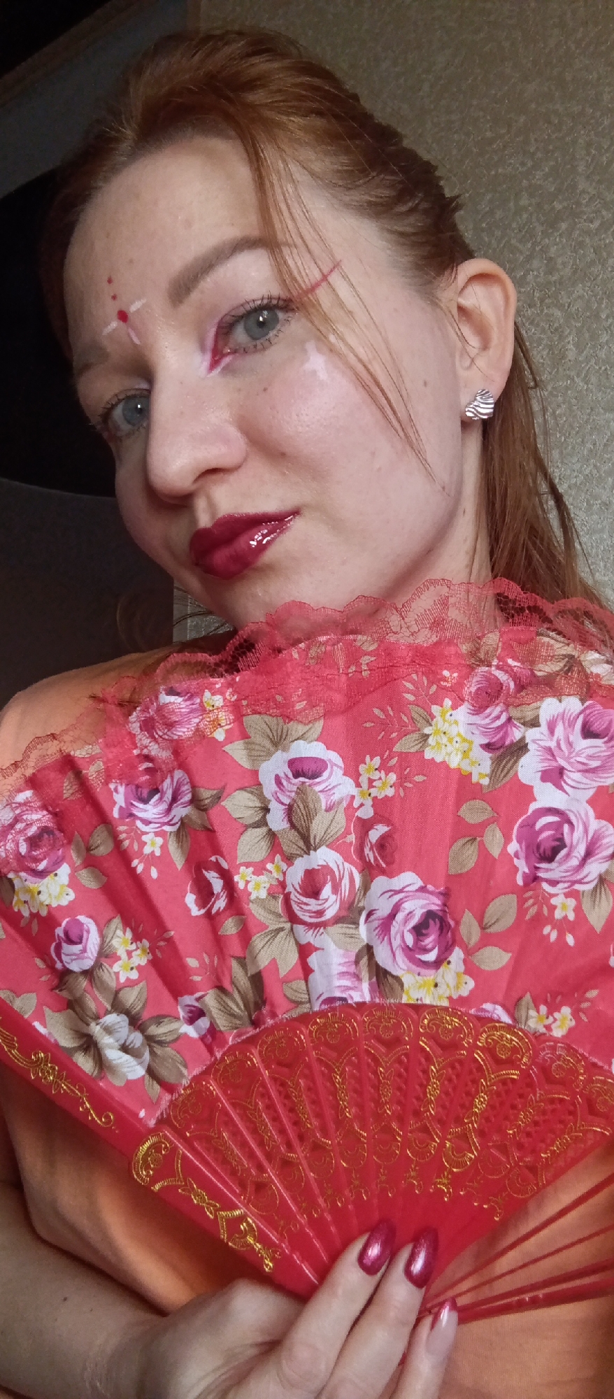 A head-and-shoulders portrait features a woman with reddish-brown hair and light blue eyes, looking directly at the camera with a subtle, confident expression. Her makeup is distinctive, including glossy dark red lipstick, a bold red eyeliner extending from the outer corner of her right eye, and white and red dots arranged in a cross-like pattern on her forehead, reminiscent of a bindi or tilak. White smudges are also visible on her left cheek and under her left eye.
She is holding a decorative red folding fan in her left hand, which partially covers her lower chest and right shoulder. The fan has a red fabric panel adorned with a vibrant floral pattern of pink and white roses, small yellow flowers, and green leaves. The fan's frame is red with intricate gold-colored designs, and a ruffled red lace trim borders the top edge of the fabric. Her fingernails are painted with a metallic reddish-pink polish. In her right ear, she wears a silver-toned stud earring with a textured, wavy design.
She is wearing an orange or coral-colored top, visible beneath and around the fan. The background is a plain, light brown or beige wall, indicating an indoor setting. The lighting is soft and even, highlighting her features and the details of the fan.