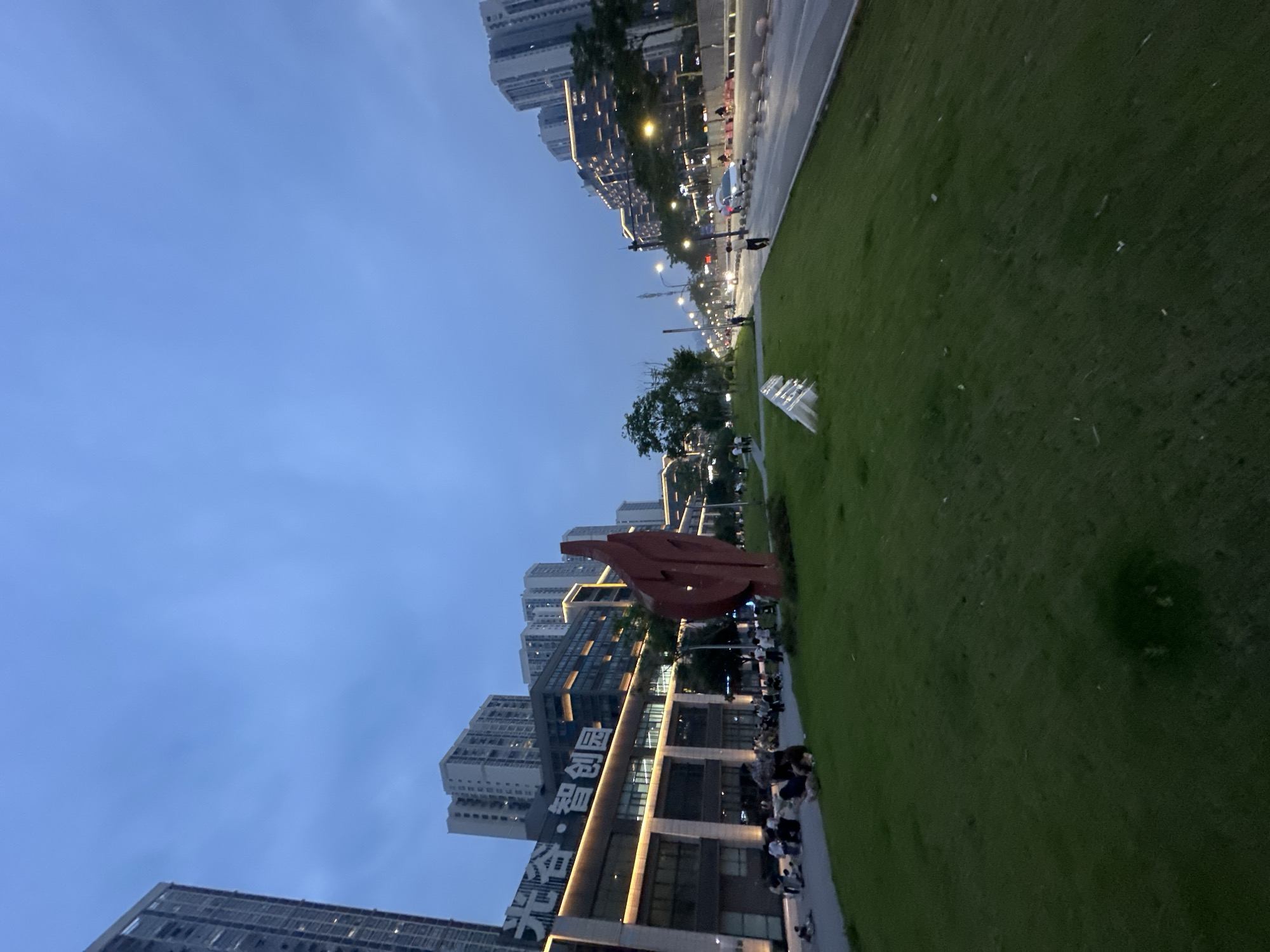 The image depicts a wide shot of a street scene in the Chongqing High-tech Zone, China, during twilight. The sky is a deep, overcast blue, hinting at the end of daylight. In the foreground, a lush green lawn slopes upwards, punctuated by a striking, abstract red sculpture resembling a stylized flame or leaf. Further back, a paved walkway or path runs alongside the lawn. Across the street, a modern multi-story building with illuminated windows and signage featuring Chinese characters ("光谷·智创园") stands as a prominent landmark. Beyond this building, a row of taller residential or office buildings stretches into the distance, their windows also lit. The street itself is active with vehicular traffic, including cars and what appear to be taxis, their headlights and taillights visible. Pedestrians are scattered along the sidewalk and on the lawn. Several individuals and small groups are seated on the grass, suggesting a relaxed atmosphere, possibly an evening gathering or a pause from urban activity. The overall impression is one of a modern, developing urban environment at dusk, with a blend of commercial and residential structures, green spaces, and ongoing city life.