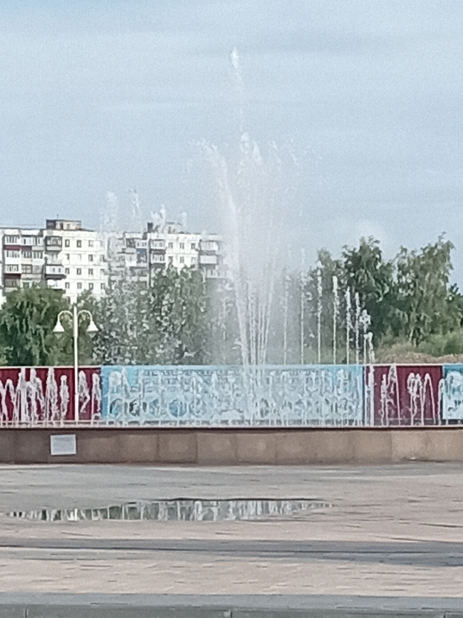 The image depicts a public fountain in Rudny, Kazakhstan. The fountain is in the foreground, with water spraying upwards and outwards. Behind the fountain, there is a decorative wall with red and blue panels. In the background, there are several multi-story apartment buildings and trees. The sky is overcast. The ground in front of the fountain is wet, with puddles reflecting the buildings. A lamppost is visible to the left of the fountain.